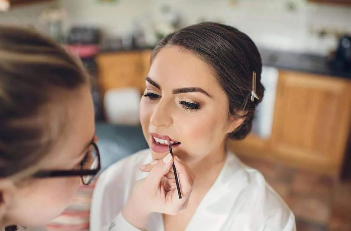 A woman getting her makeup done, with a makeup artist applying lipstick with a brush.
