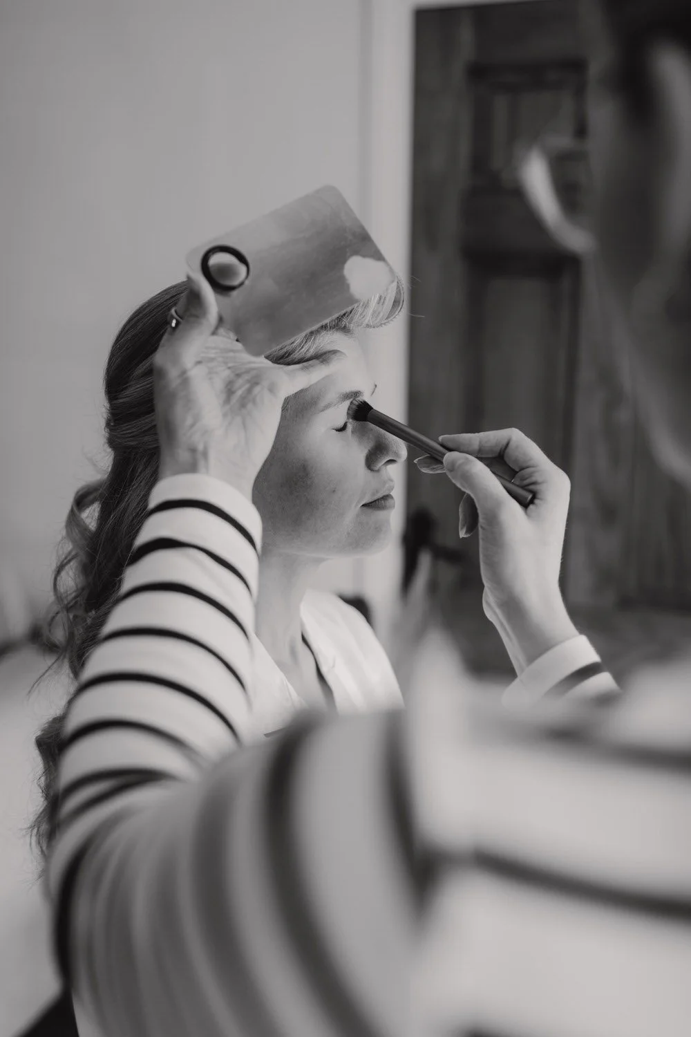 A woman with closed eyes is having makeup applied by a makeup artist, who is using a brush near her eyelid. The artist is holding a small mirror or makeup palette above her head. The woman has wavy hair and is wearing a striped top.