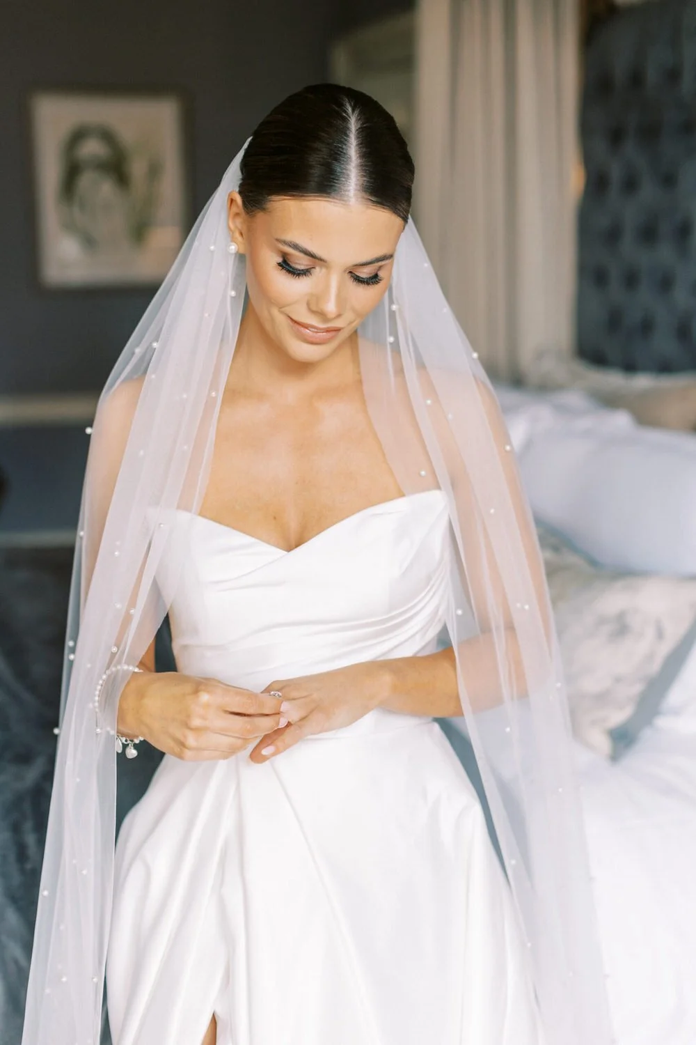 A bride in a white wedding dress and veil, looking down and smiling softly, in a softly lit interior room.
