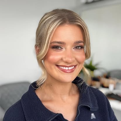 Close-up of a young woman with blonde hair, smiling, wearing a dark collared top, in a modern indoor setting.