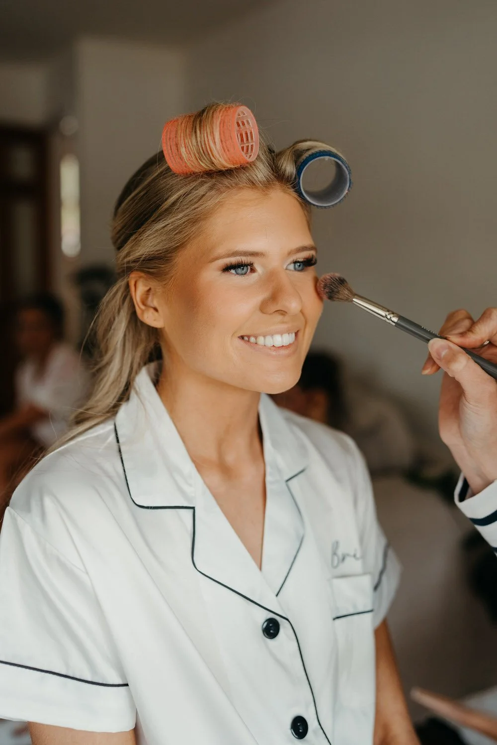 A woman with hair rollers smiling while a makeup artist applies blush to her cheek.
