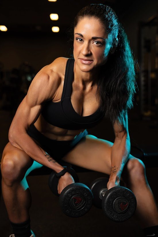 A fit woman with long dark hair working out with dumbbells in a gym.