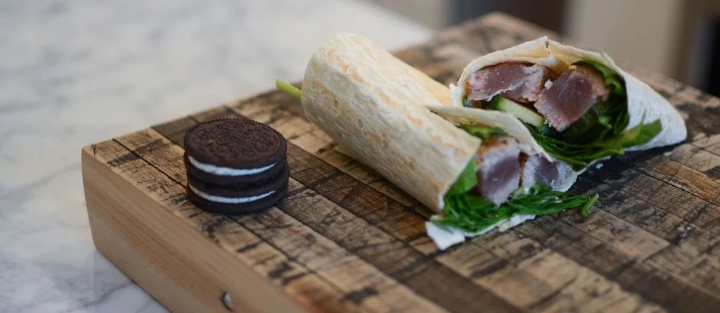 A wrap sandwich with greens and meat, next to a stack of Oreo cookies on a wooden cutting board.