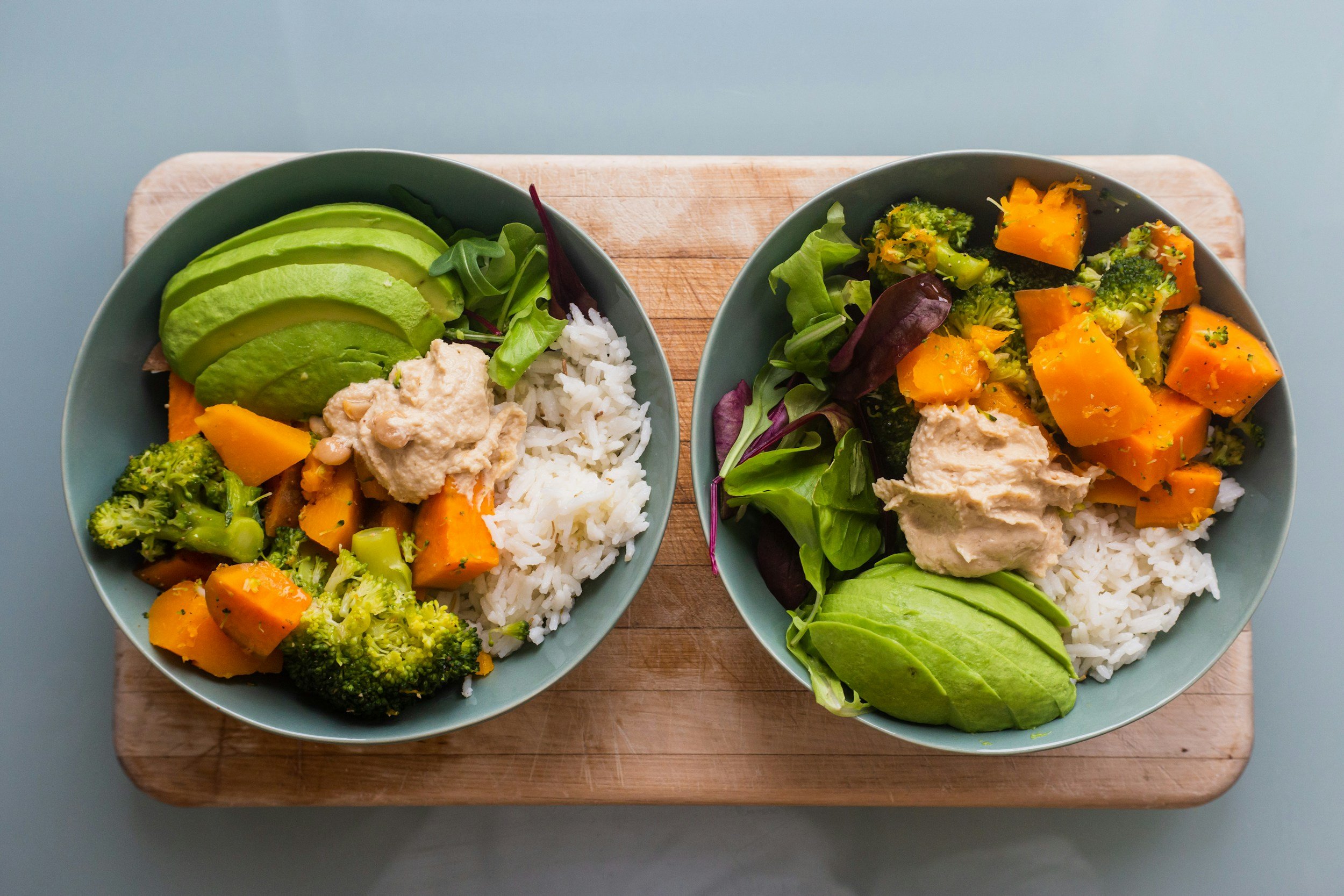 Two bowls of colorful vegetable and rice dishes on a wooden tray. Each bowl contains sliced avocado, broccoli, roasted sweet potatoes, mixed greens, and a scoop of hummus, served over white rice.
