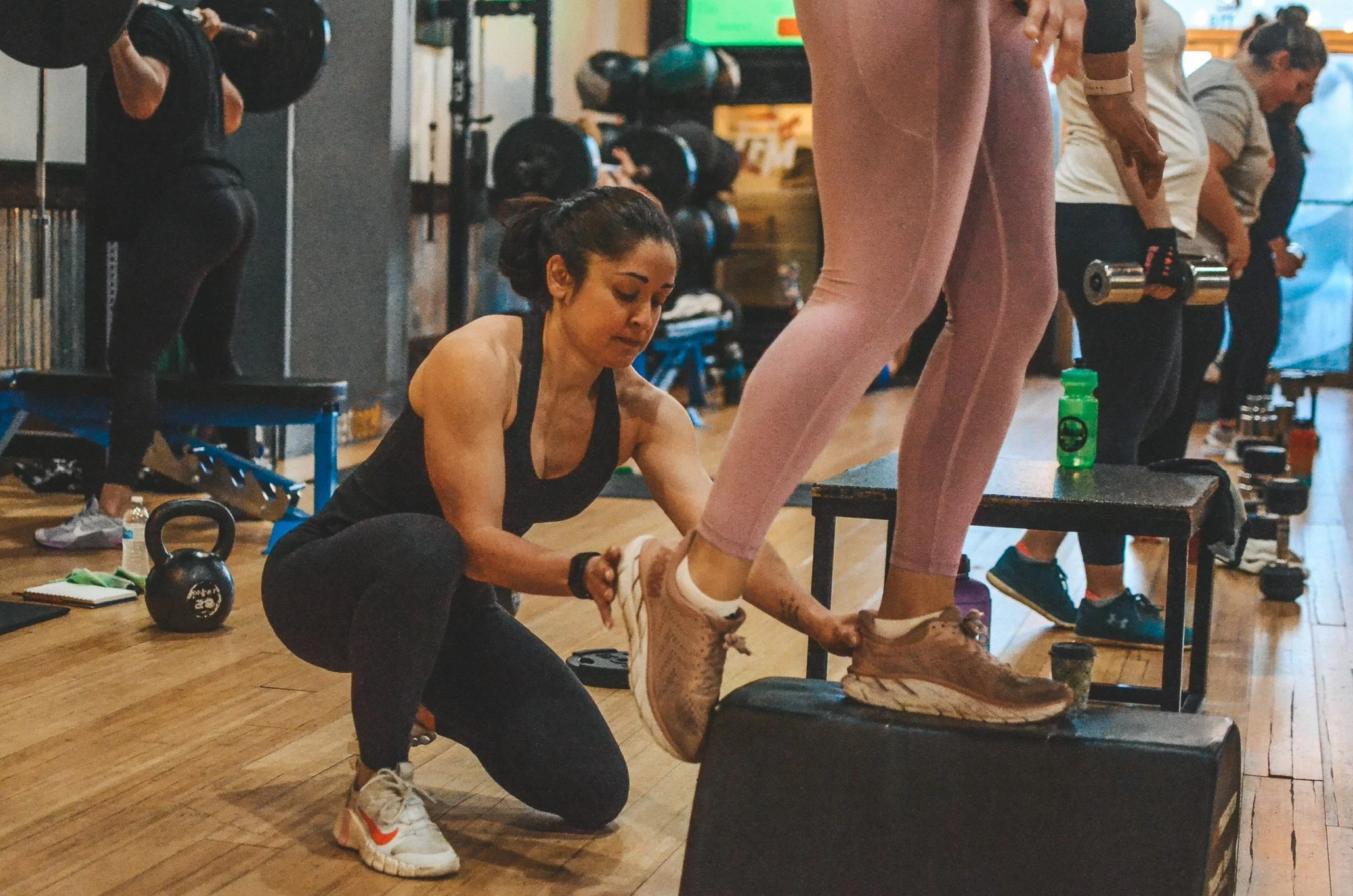 A woman is helping another woman, who is standing on a box in a gym, tighten her shoelaces. Other people are working out in the background, and fitness equipment such as dumbbells and kettlebells are visible.
