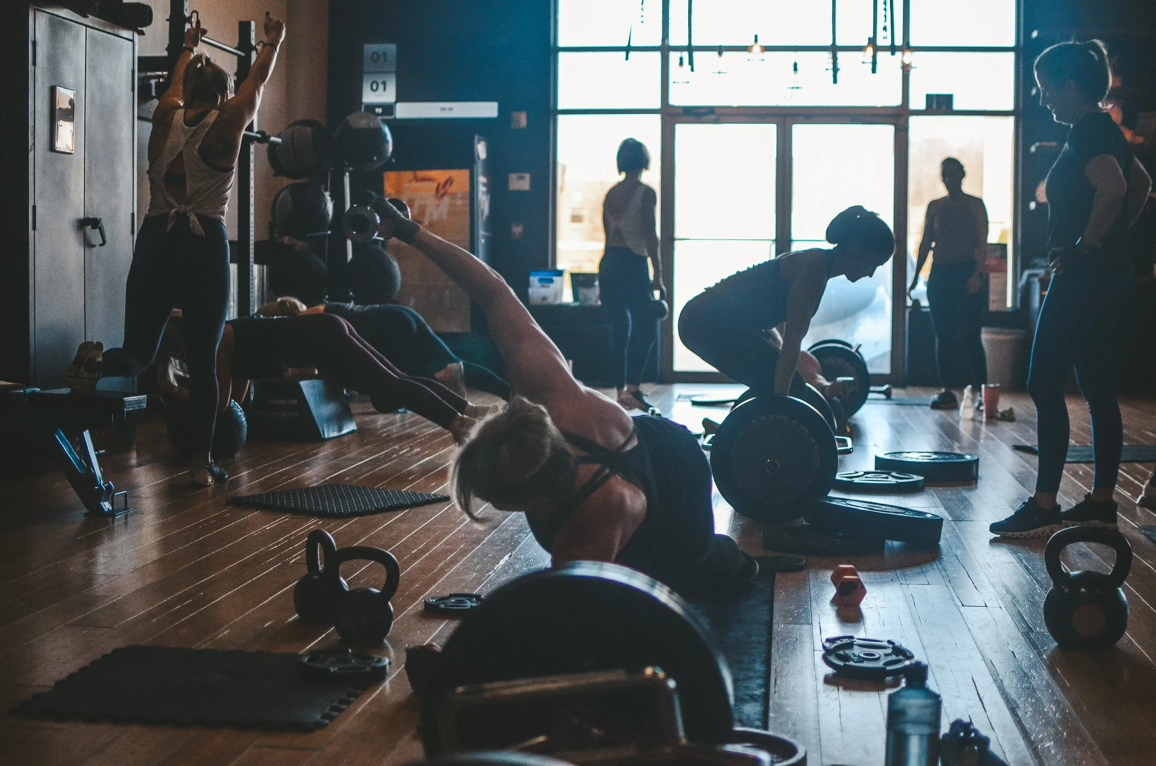 People working out in a gym, some lifting weights, others stretching or resting, with a large window in the background.