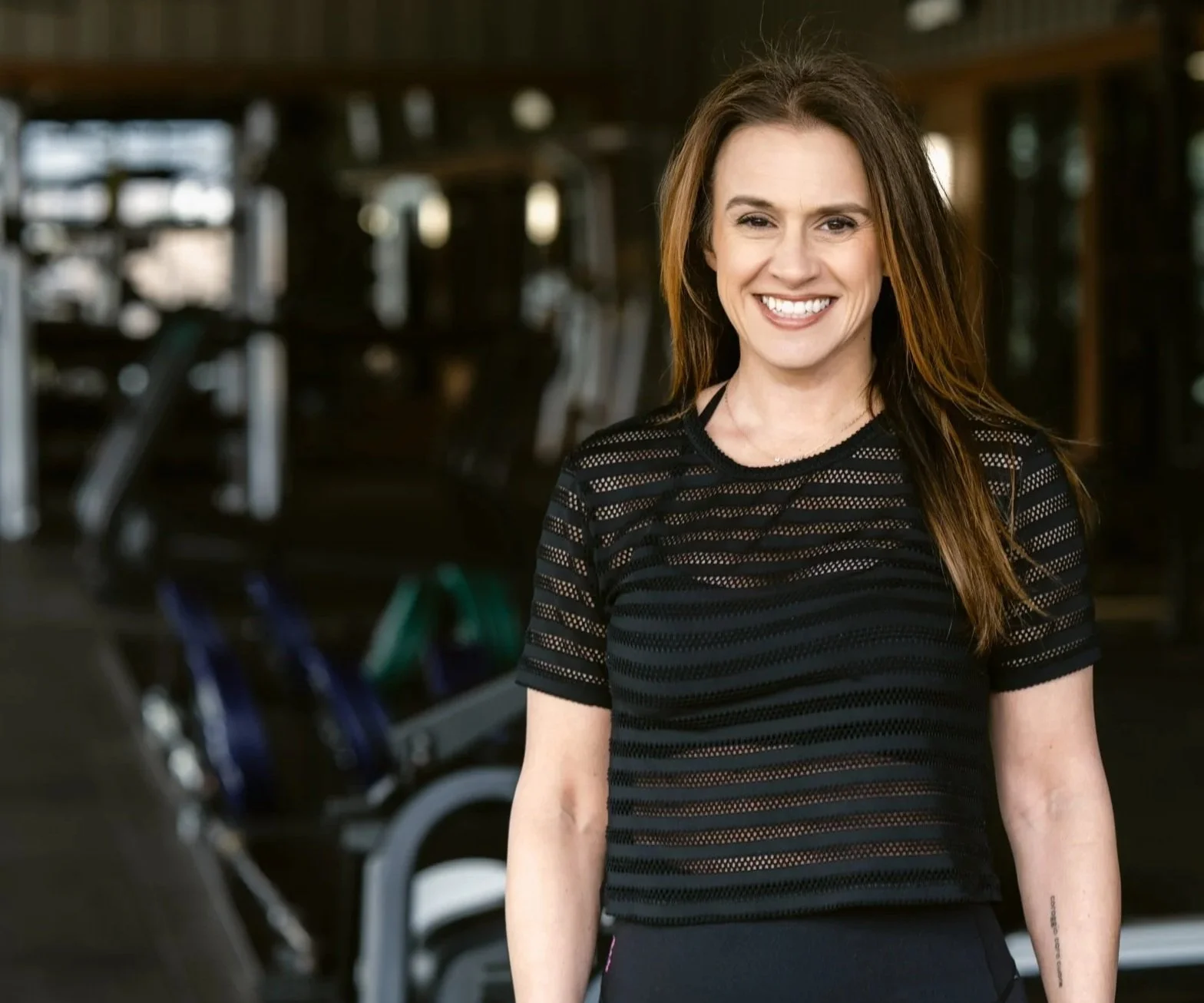 A smiling woman with long brown hair in a gym, wearing a black mesh short-sleeve top.