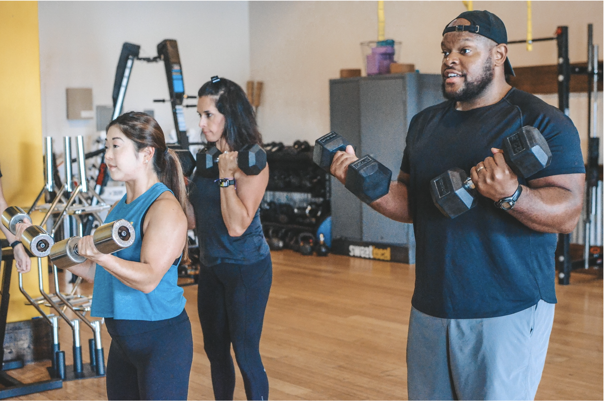 Three people lifting dumbbells during a workout in a gym, with a focus on a man with a baseball cap and dark shirt, and two women in athletic wear.