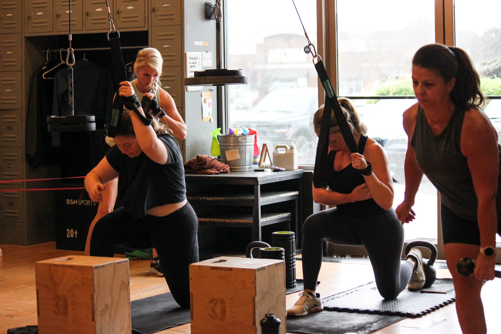 Women participating in a fitness class with one woman holding a weighted strap, others kneeling with fists clenched, surrounded by workout equipment and boxes, inside a gym with lockers and large windows.
