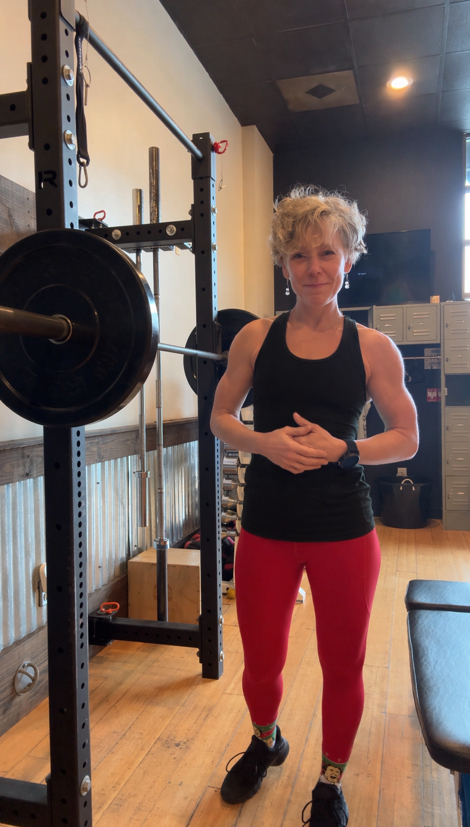 A woman with short curly blonde hair, wearing a black tank top, red leggings with colorful socks, and black shoes, standing next to a weightlifting rack in a gym.