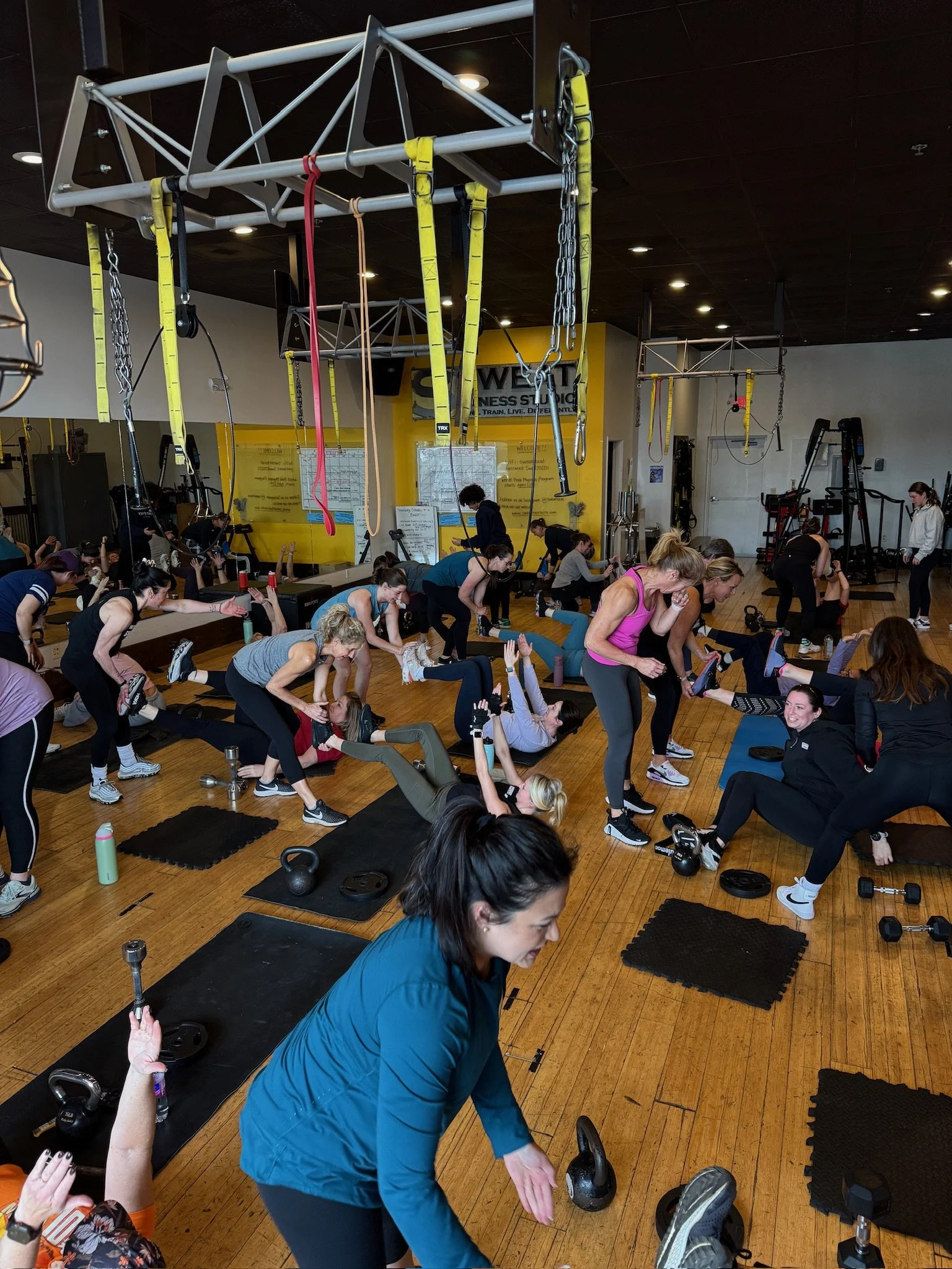 People participating in a group fitness class or workout session at a gym, with some women doing exercises with kettlebells and others engaging in stretching or using resistance bands, in a well-lit gym that has various fitness equipment and a yellow wall with a logo.