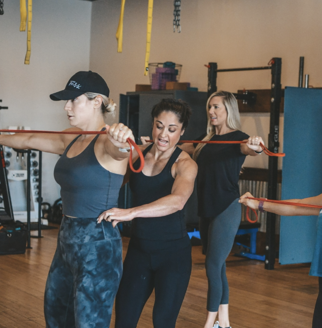 Three women participating in a fitness class using resistance bands for arm exercises in a gym.