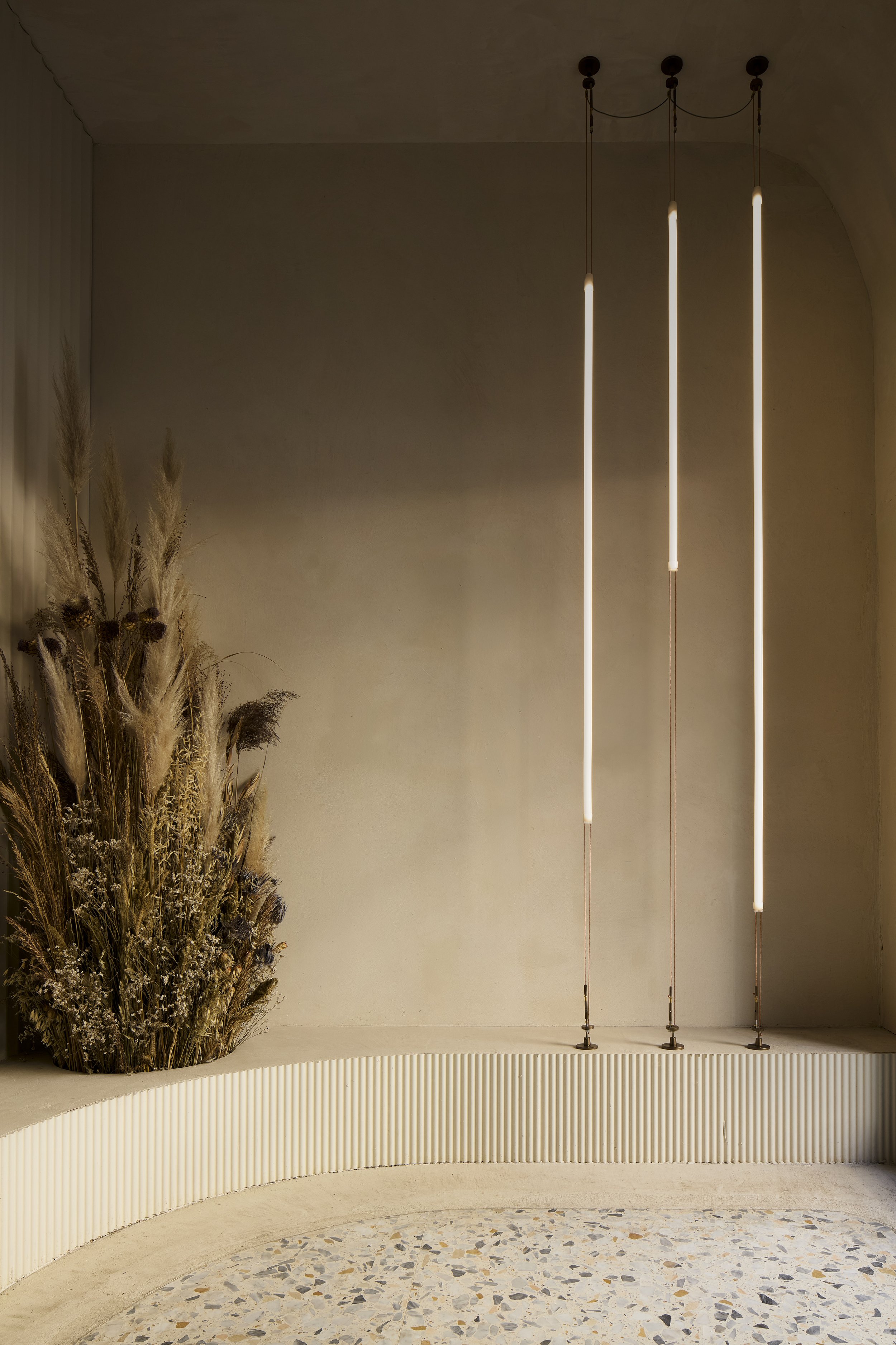 Interno minimalista con bouquet di fiori secchi e luci pendenti lungo una parete beige, con soffitto arcuato e pavimento in pietra multicolore.