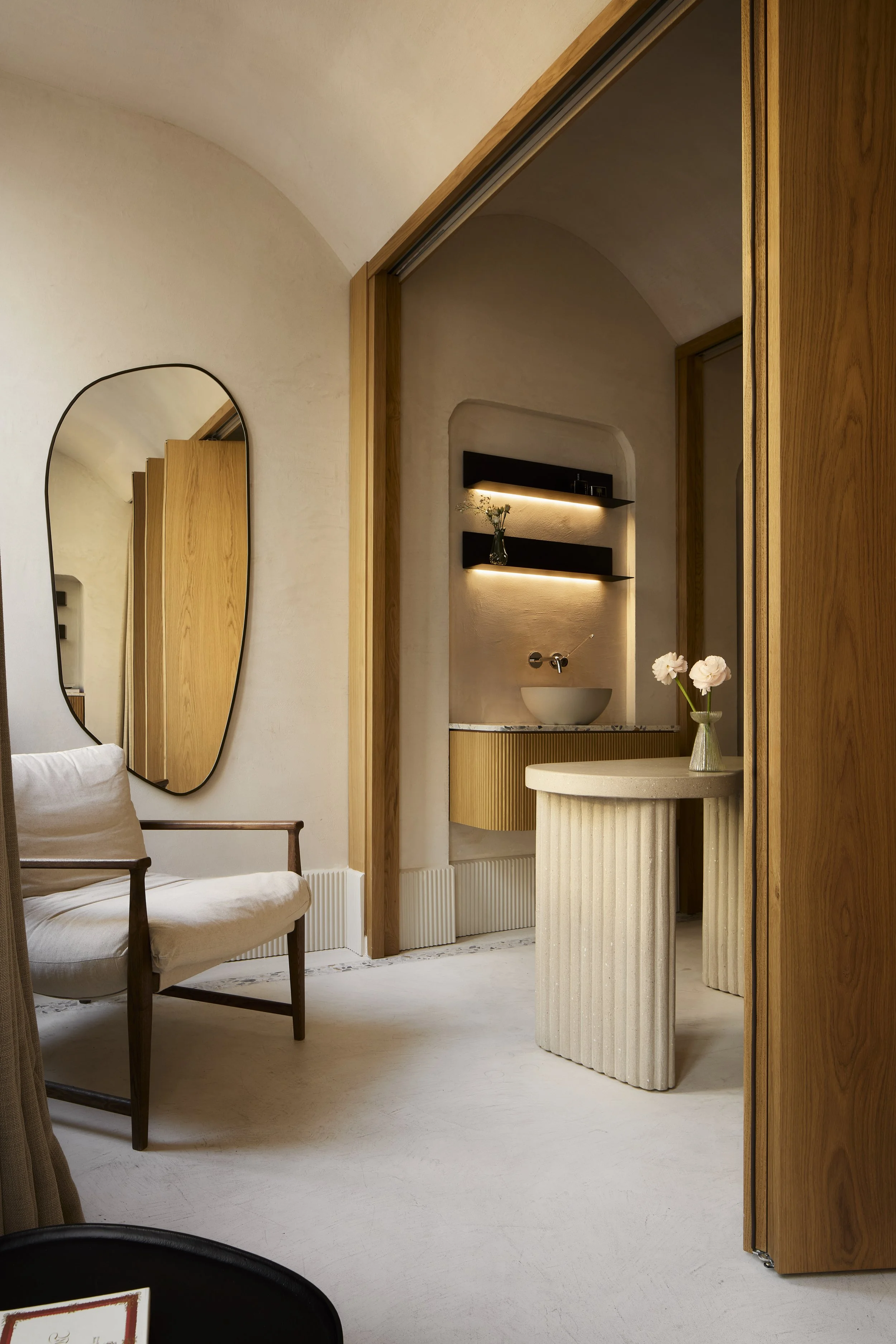 Interno di una stanza con un mobile in legno, un grande specchio a forma irregolare sulla parete e un tavolo con vasi di fiori, pareti di colore beige e un angolo con lavabo e scaffali illuminati.