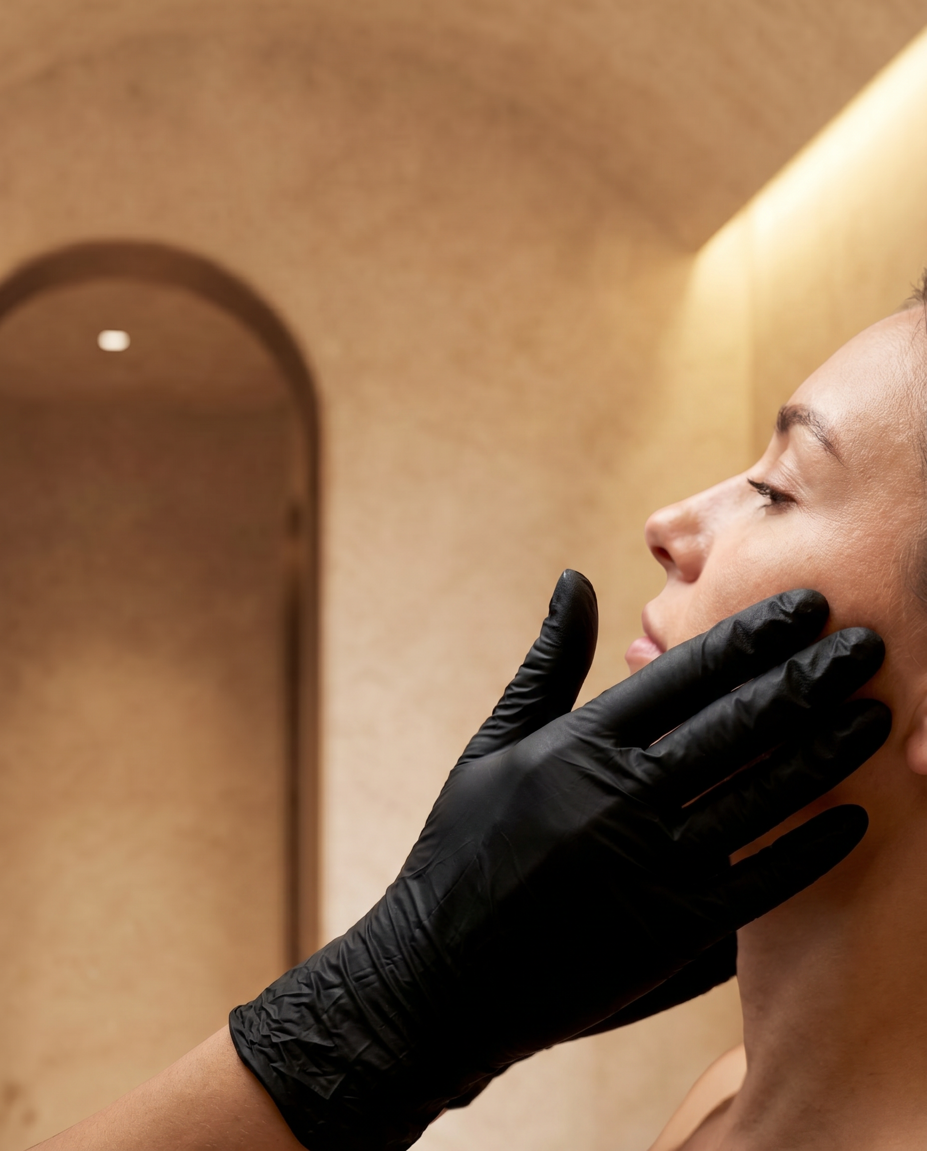 Prima di un trattamento di bellezza, una donna con la pelle liscia e senza trucco riceve una seduta di cura del viso da una professionista con guanti neri, al centro di un ambiente calmo e ben illuminato.