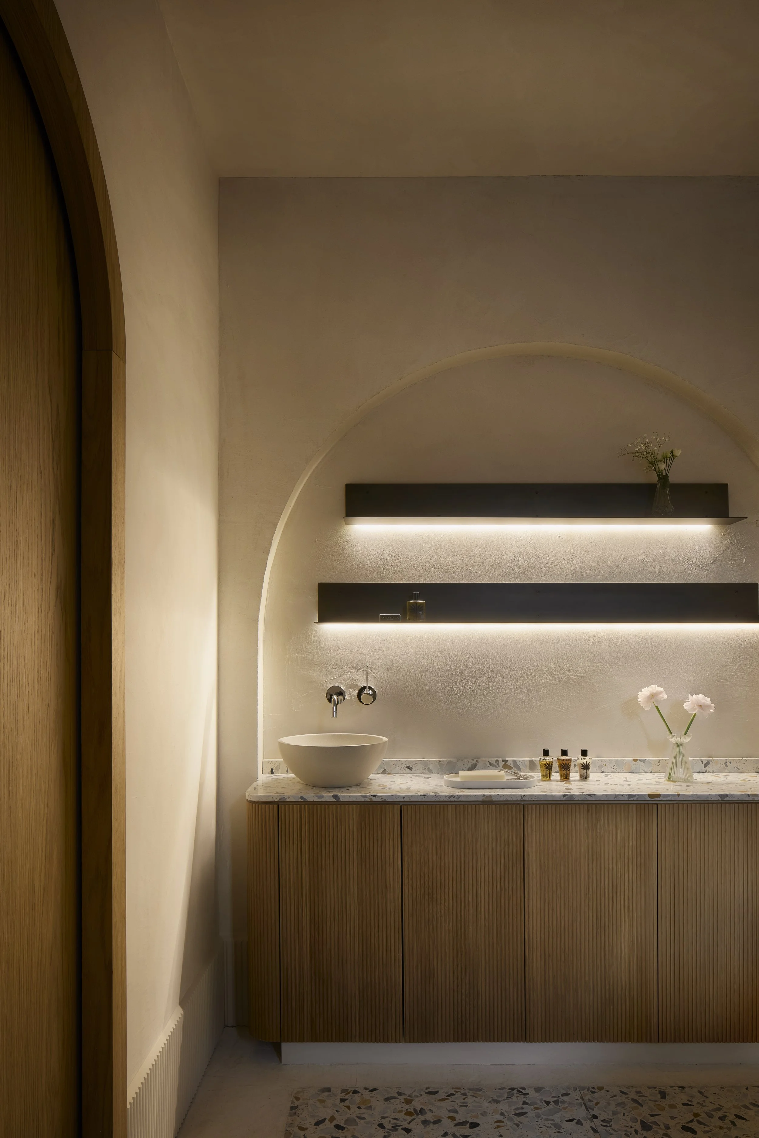 Barra a muro in stile minimalista con lavabo rotondo bianco sul piano in marmo, mensole nere con piccole bottiglie di oli essenziali o cosmetici, e un vaso con fiori chiari. Pareti e soffitto in tonalità beige, illuminazione morbida.