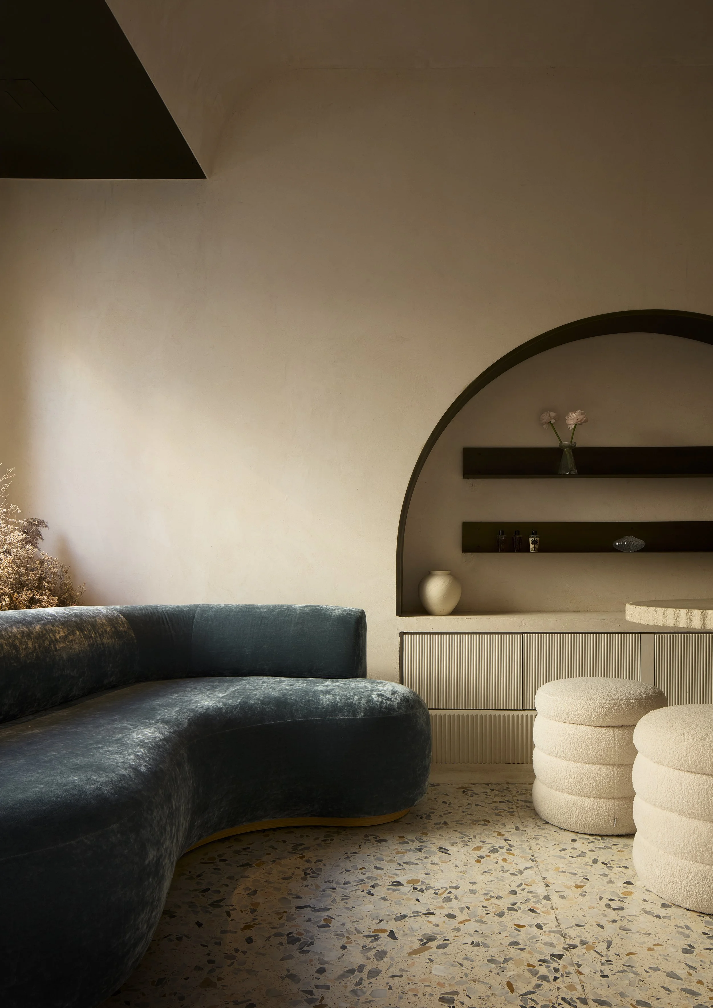 Interno di una stanza con divano blu scuro, pareti beige, scaffali neri con vasi e decorazioni, e due sedute bianche a forma di cilindro, con pavimento in mosaico