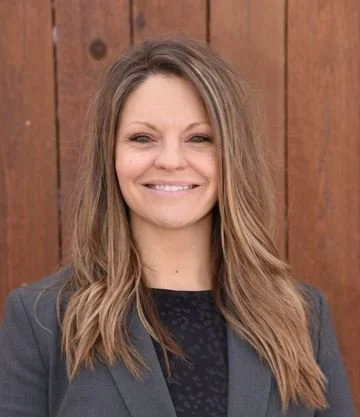A smiling woman with shoulder-length light brown hair, wearing a dark blazer, standing in front of a wooden background.