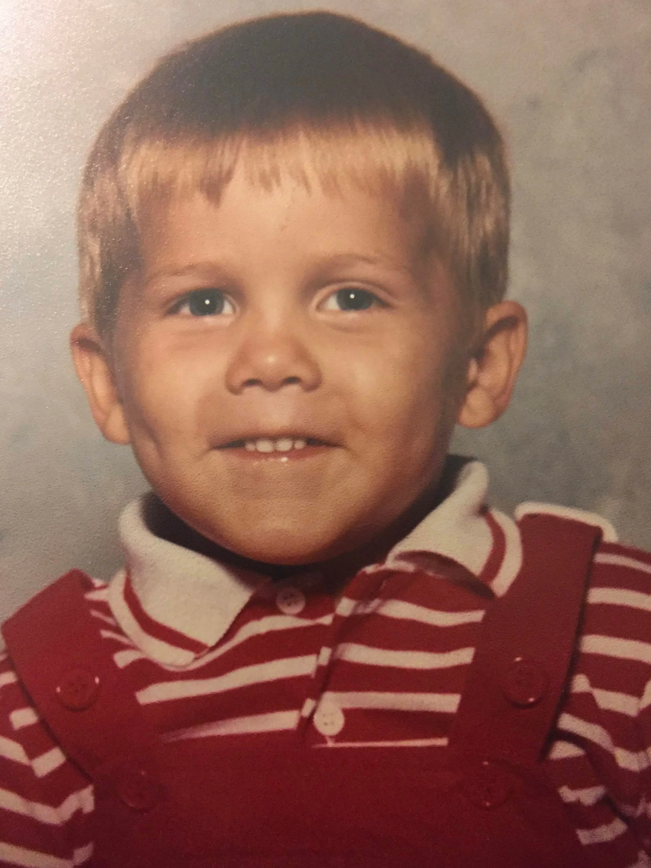 A young boy with short brown hair and blue eyes smiling, wearing a red and white striped shirt with buttons.
