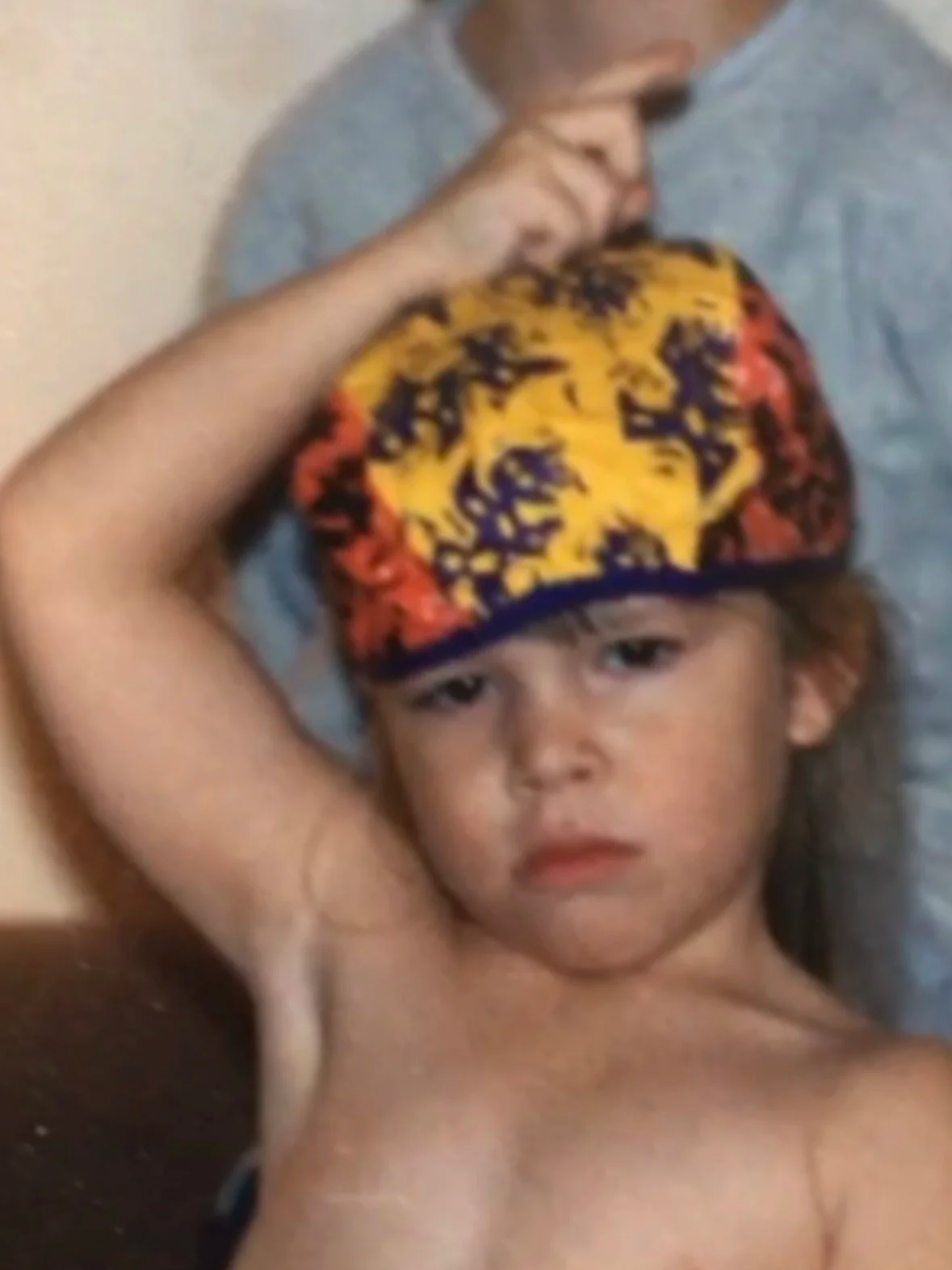 A young boy with long hair wearing a colorful hat with red, yellow, and black patterns, looking at the camera with a serious expression.