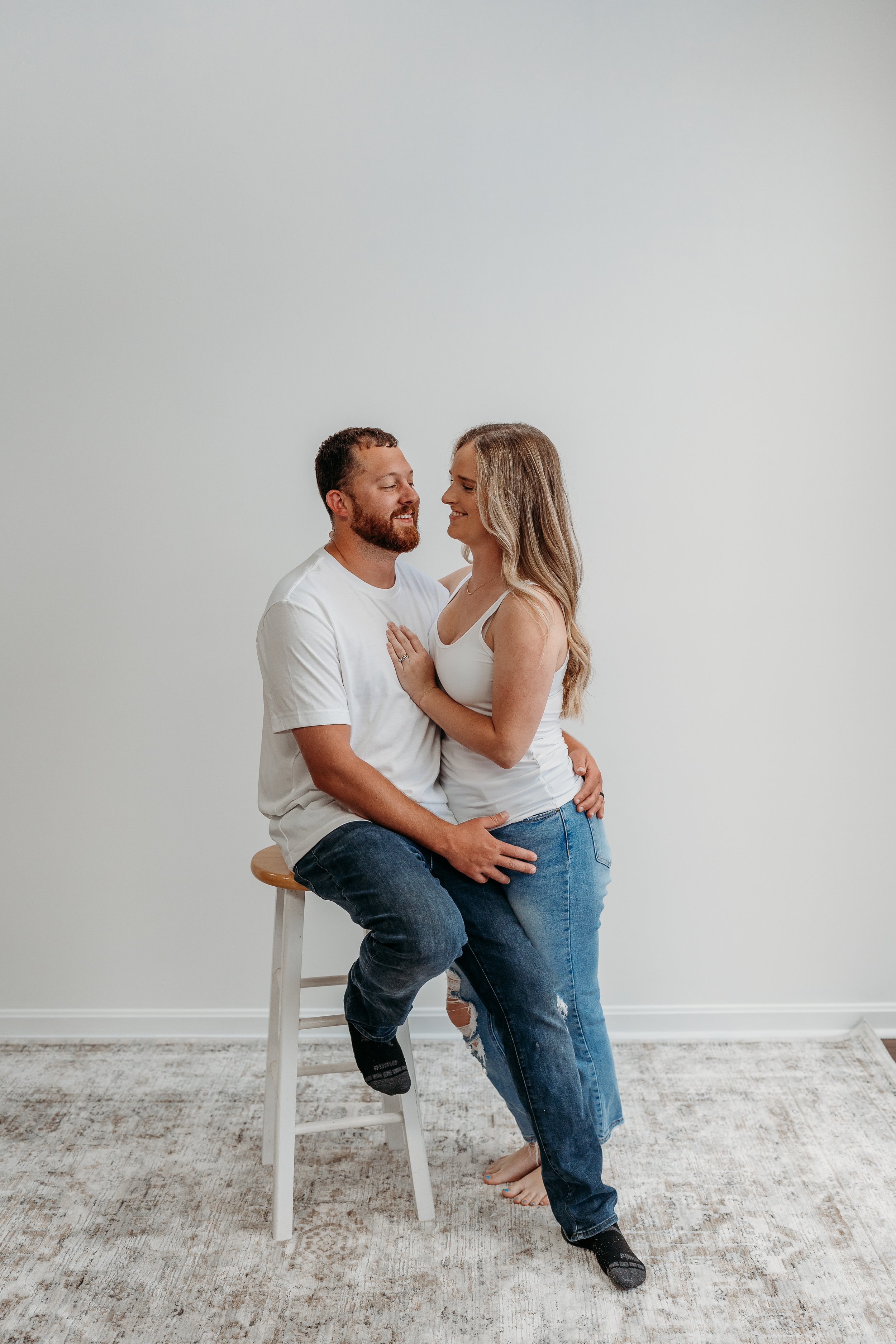 A couple sitting and standing closely together, smiling and looking at each other, in an indoor setting with a plain white wall background.