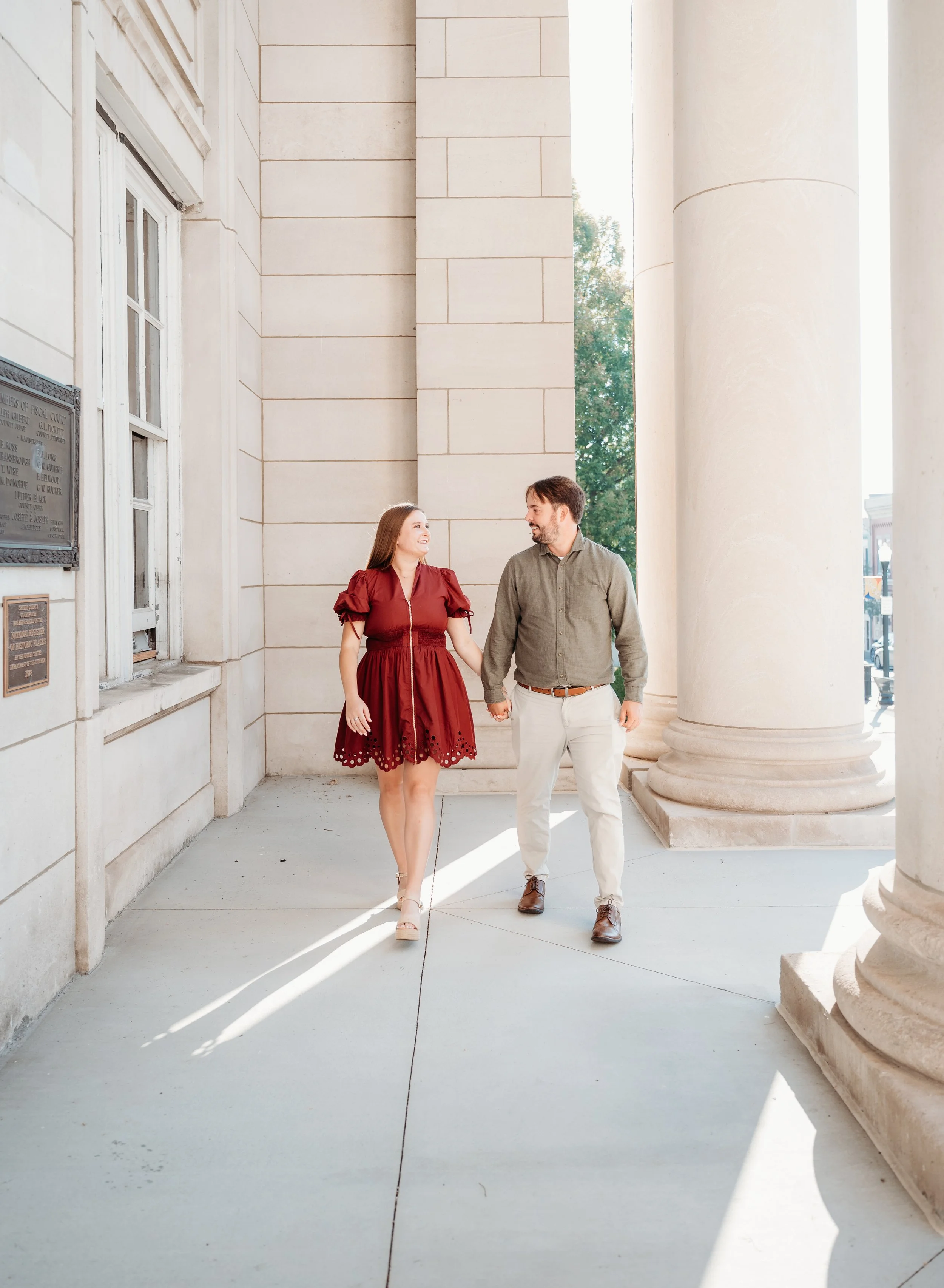 A young woman and a young man are walking hand in hand outside a neoclassical building with large columns. The woman is wearing a red dress with puffed sleeves and beige high-heeled shoes. The man is dressed in a light gray button-up shirt, white pants, and brown shoes. They are smiling and looking at each other.
