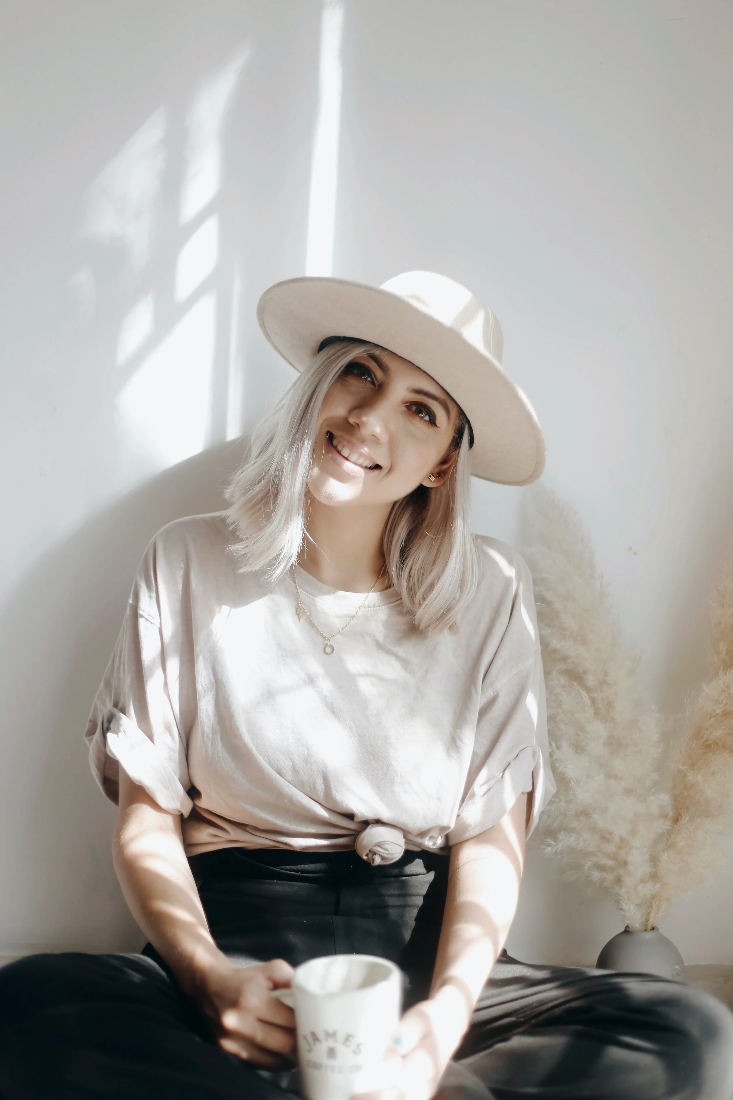 A young woman with blonde hair wearing a wide-brimmed hat, sitting against a white wall, holding a mug, smiling at the camera, with natural sunlight casting shadows on the wall.