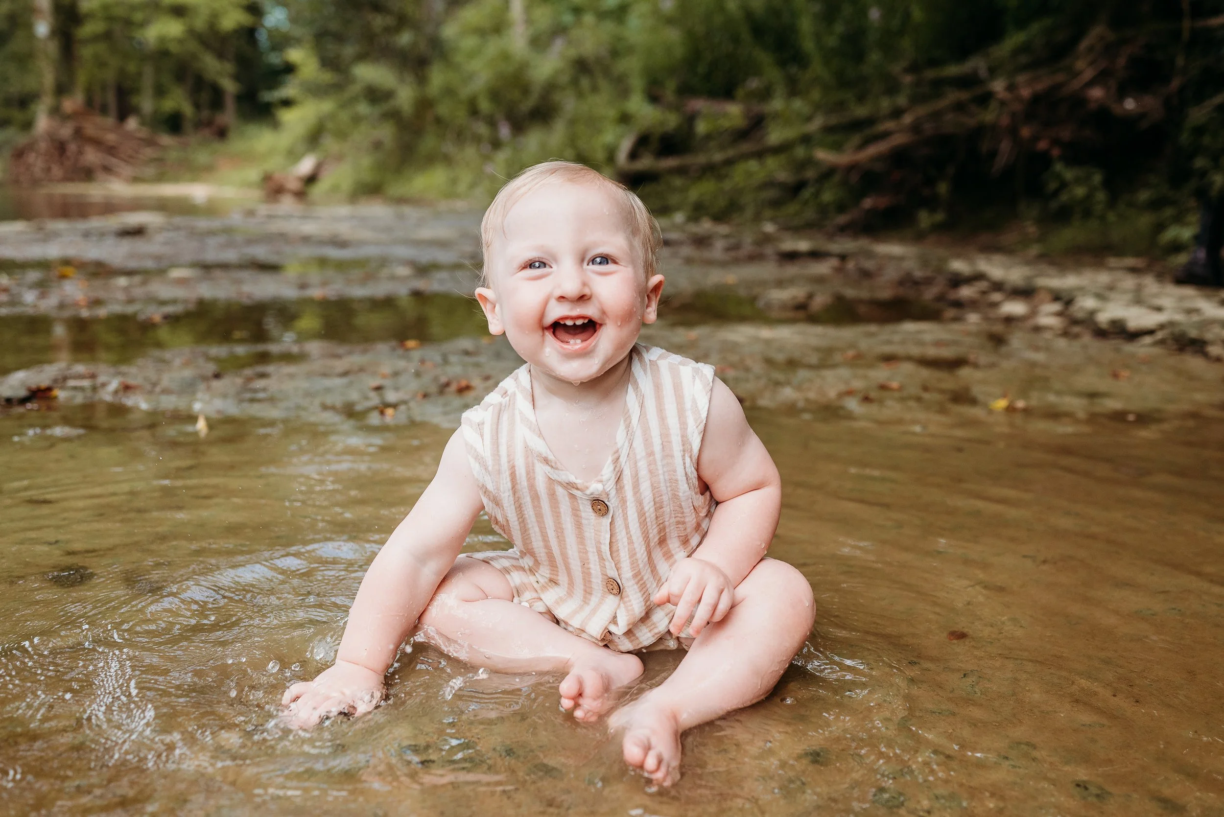 A smiling baby sitting in a shallow creek with a forest background.