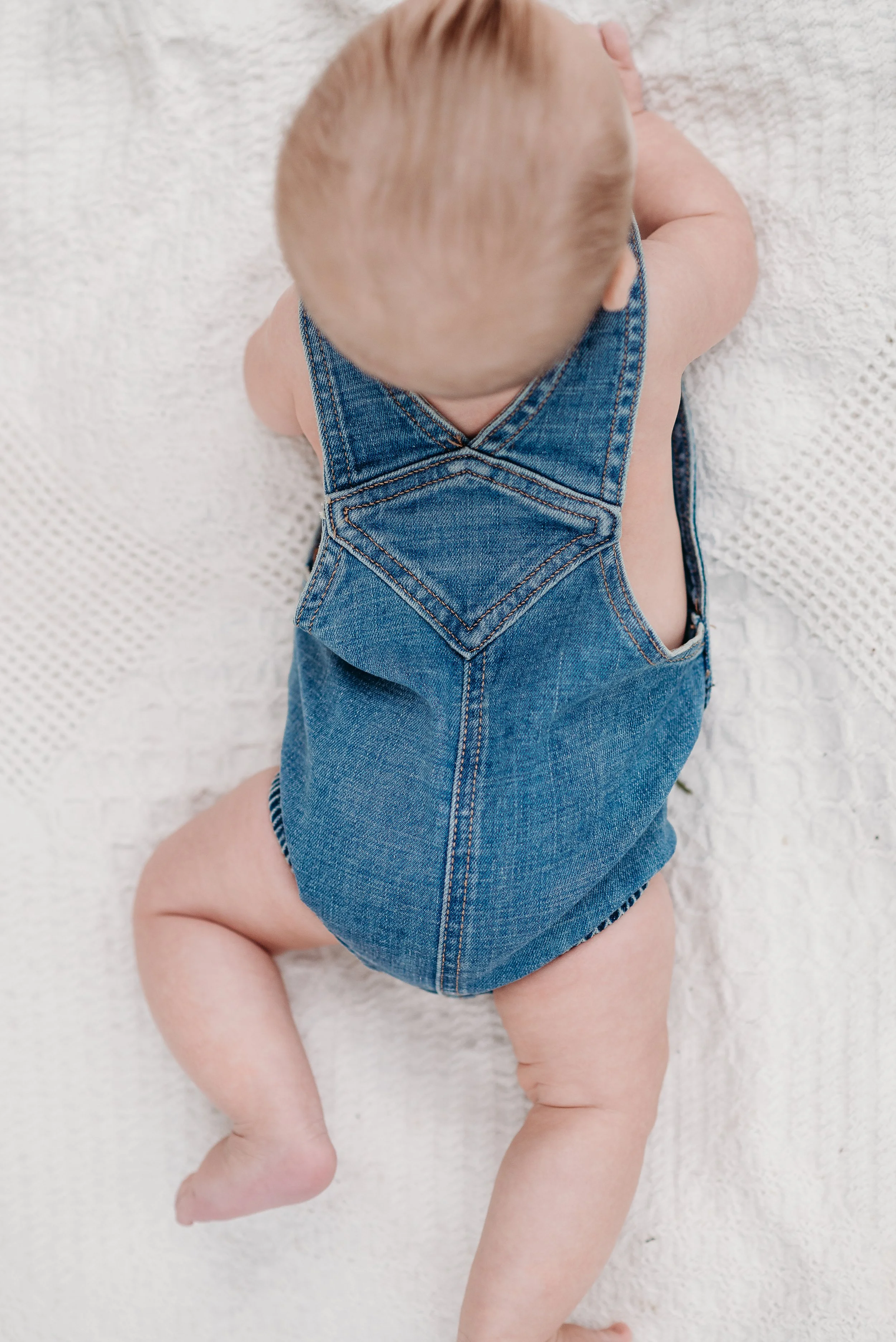 A baby wearing a denim overall crawling on a white textured blanket.