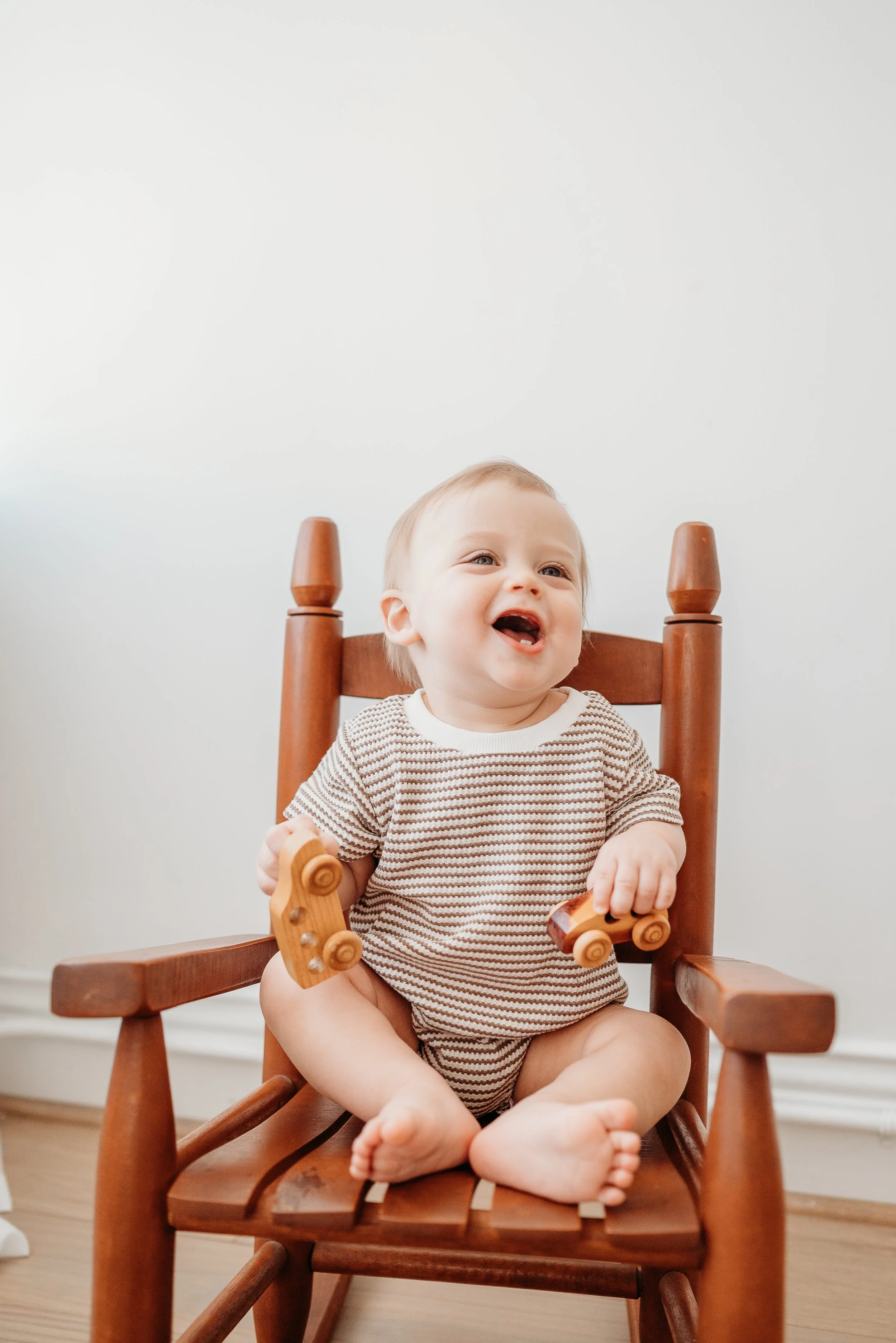 A smiling toddler sitting cross-legged on a wooden rocking chair, holding wooden toy vehicles, with a white wall background.
