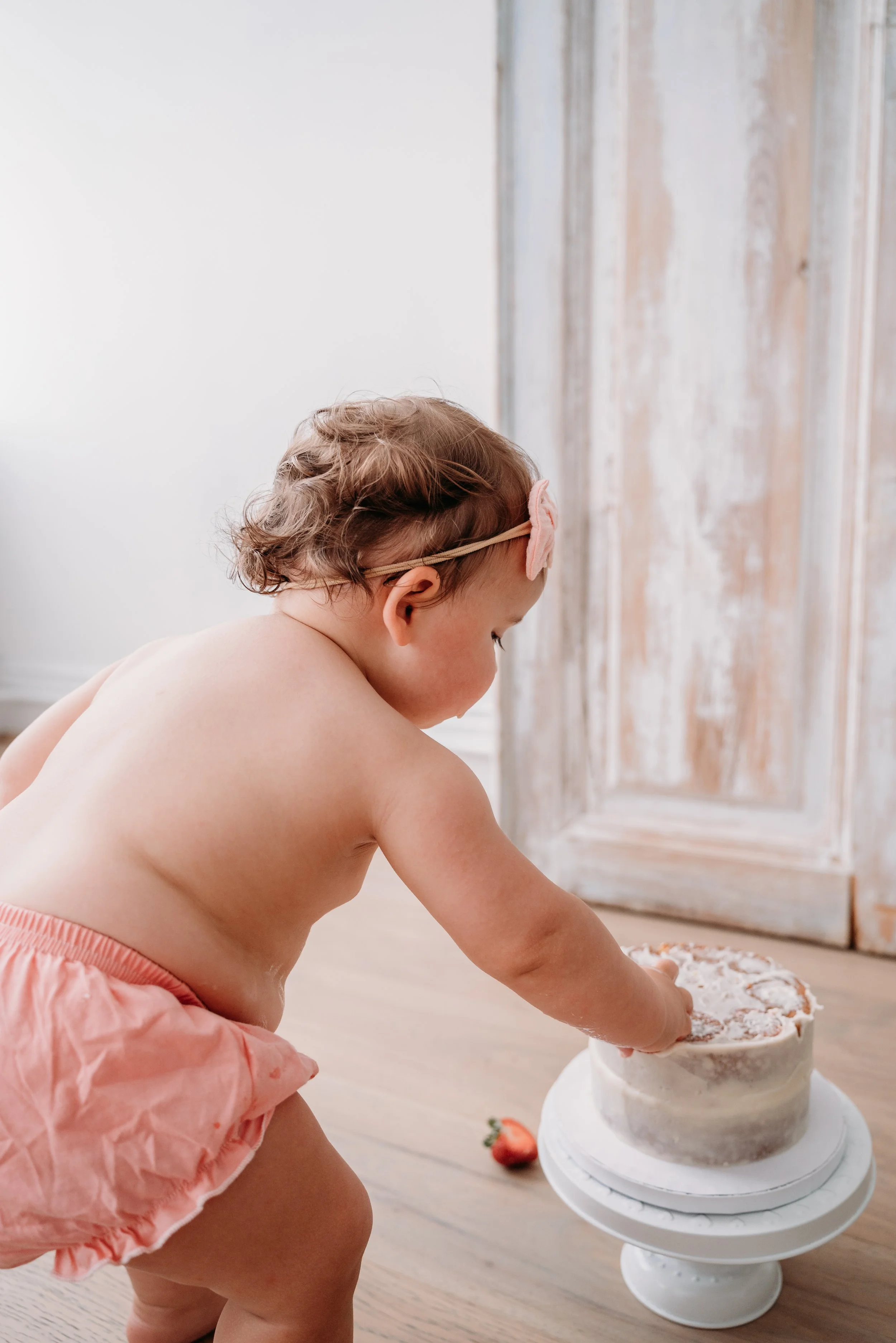 A young girl, wearing a pink bow headband and pink dress, reaches for a cake on a white stand in a room with wooden floors and a rustic door.