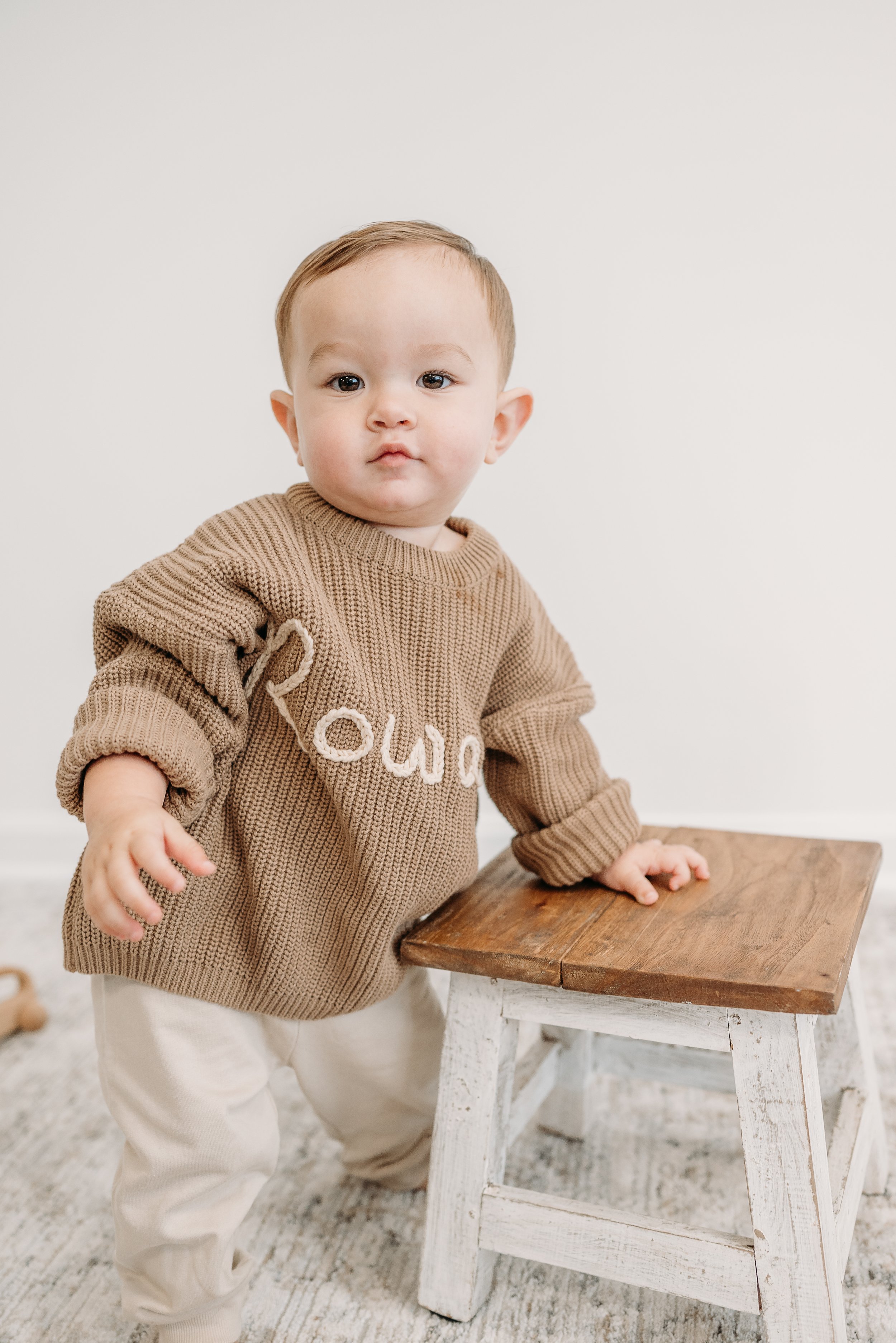 A young child with light skin and short hair standing next to a small wooden stool, looking at the camera with a neutral expression, wearing a brown sweater and light-colored pants.