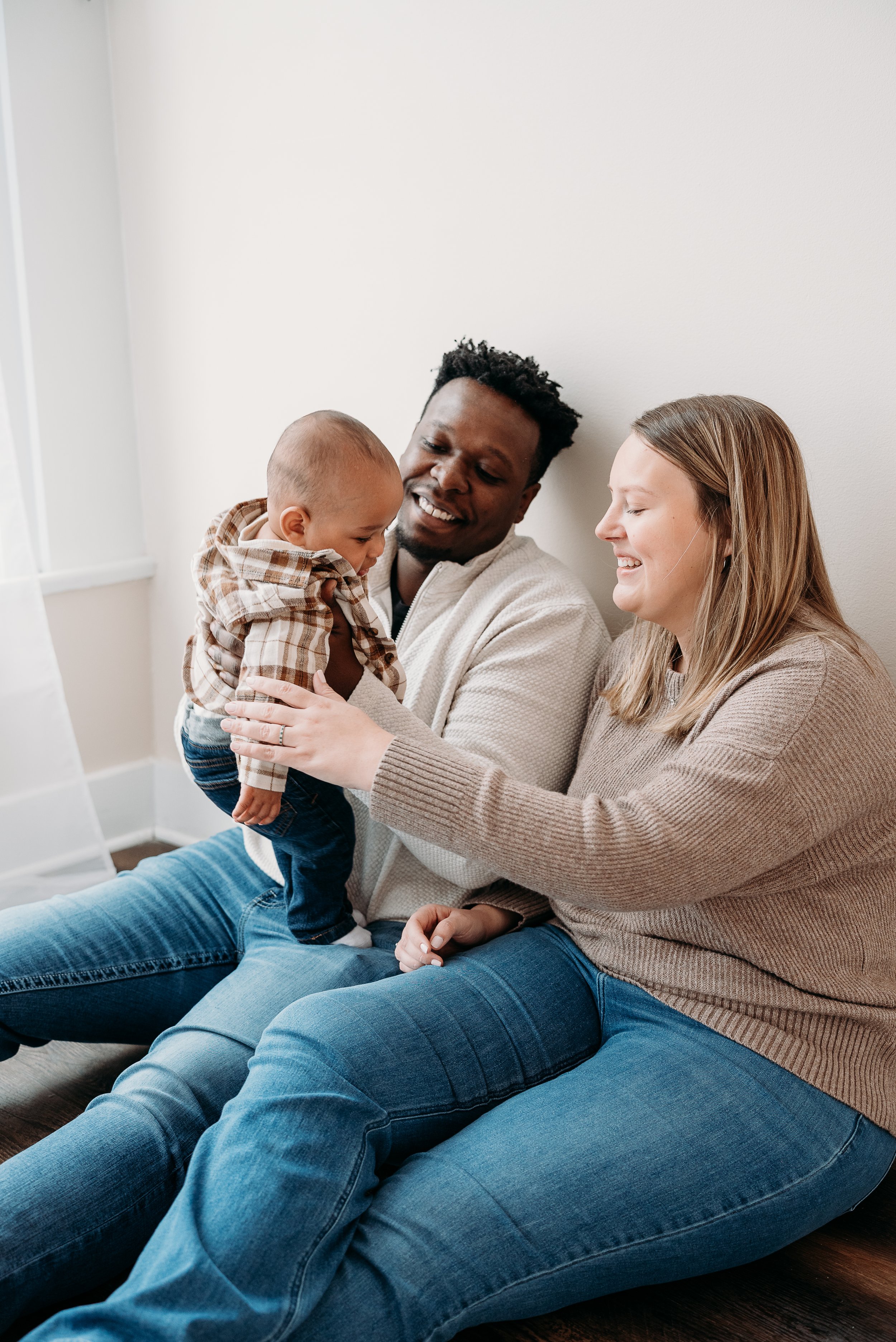 A happy family sitting on the floor, playing with a young child, in a cozy room with white walls.