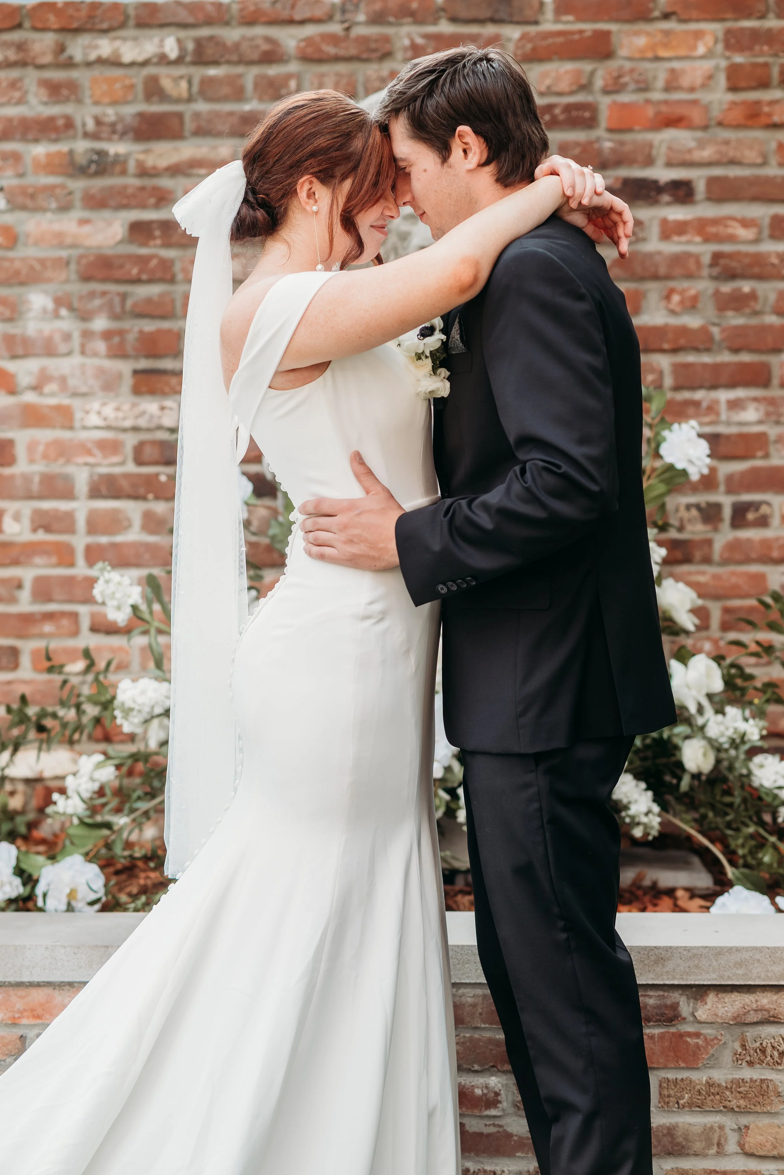 A bride and groom sharing a close, intimate moment during their wedding, with foreheads touching and arms around each other, standing in front of a brick wall with flowers.