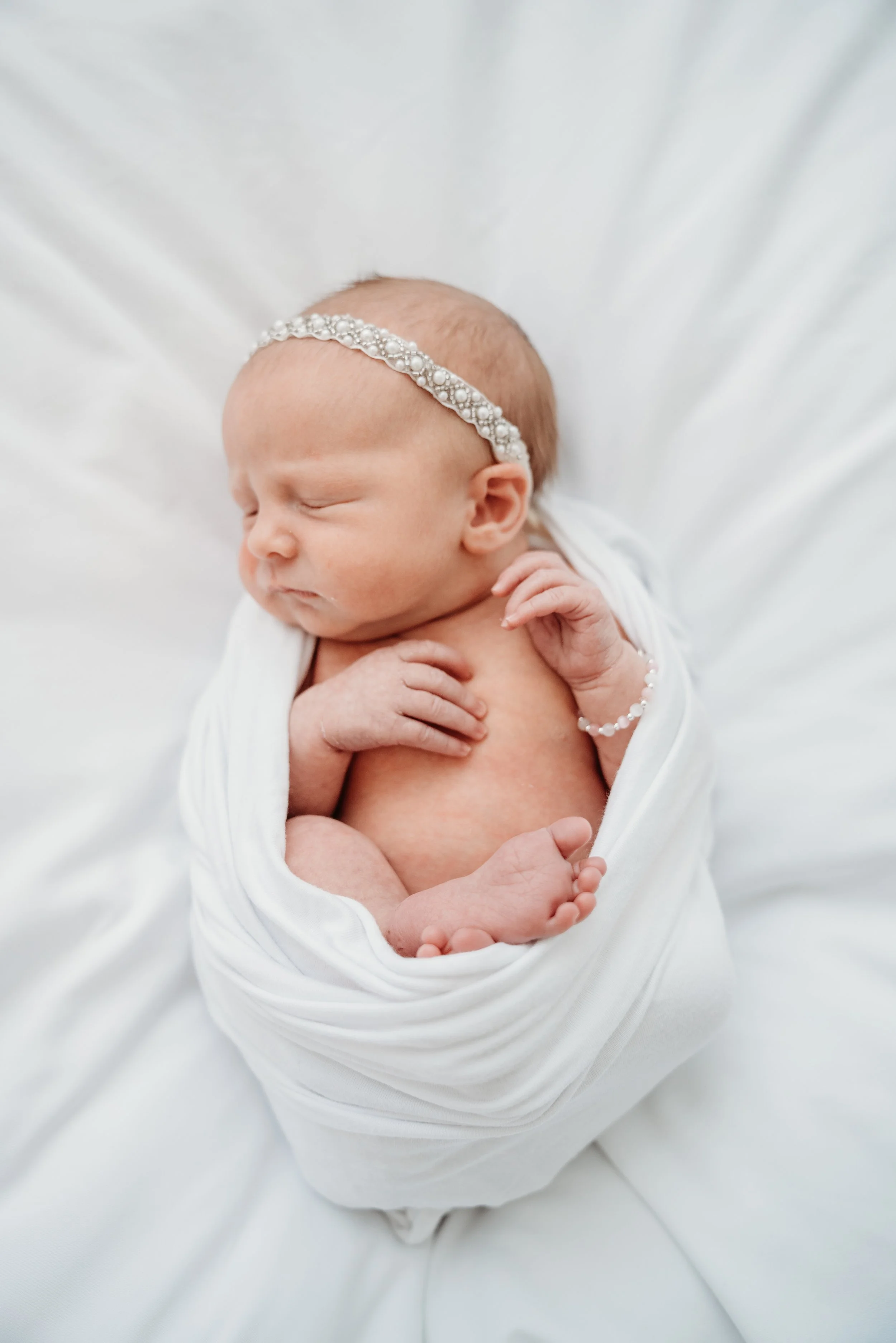A newborn baby girl wrapped in a white blanket, sleeping on a white surface, wearing a pearl headband and bracelet.