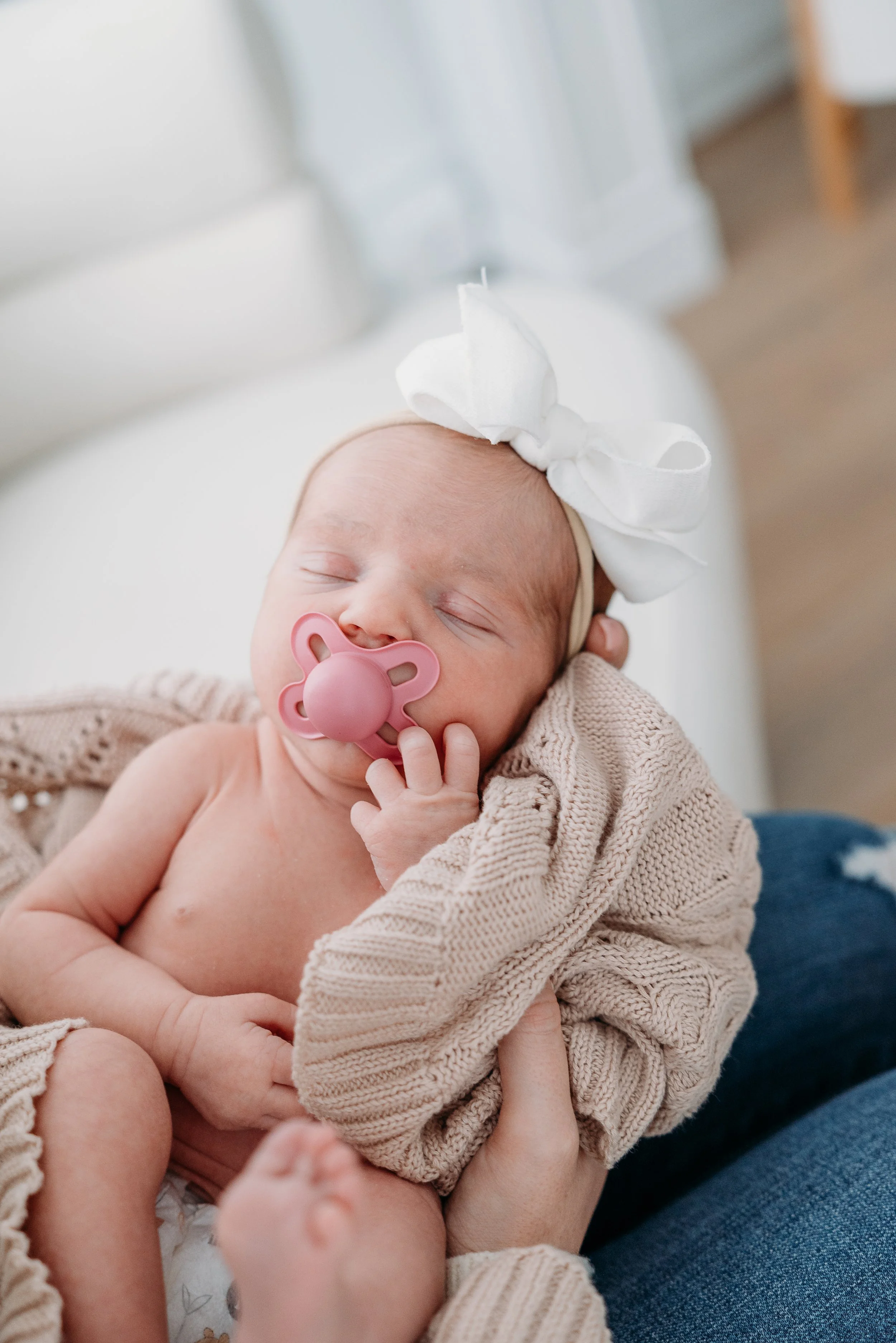 Close-up of a sleeping newborn baby with a pink pacifier and a white bow headband, held gently by an adult's hand, cozy in a beige knitted blanket.