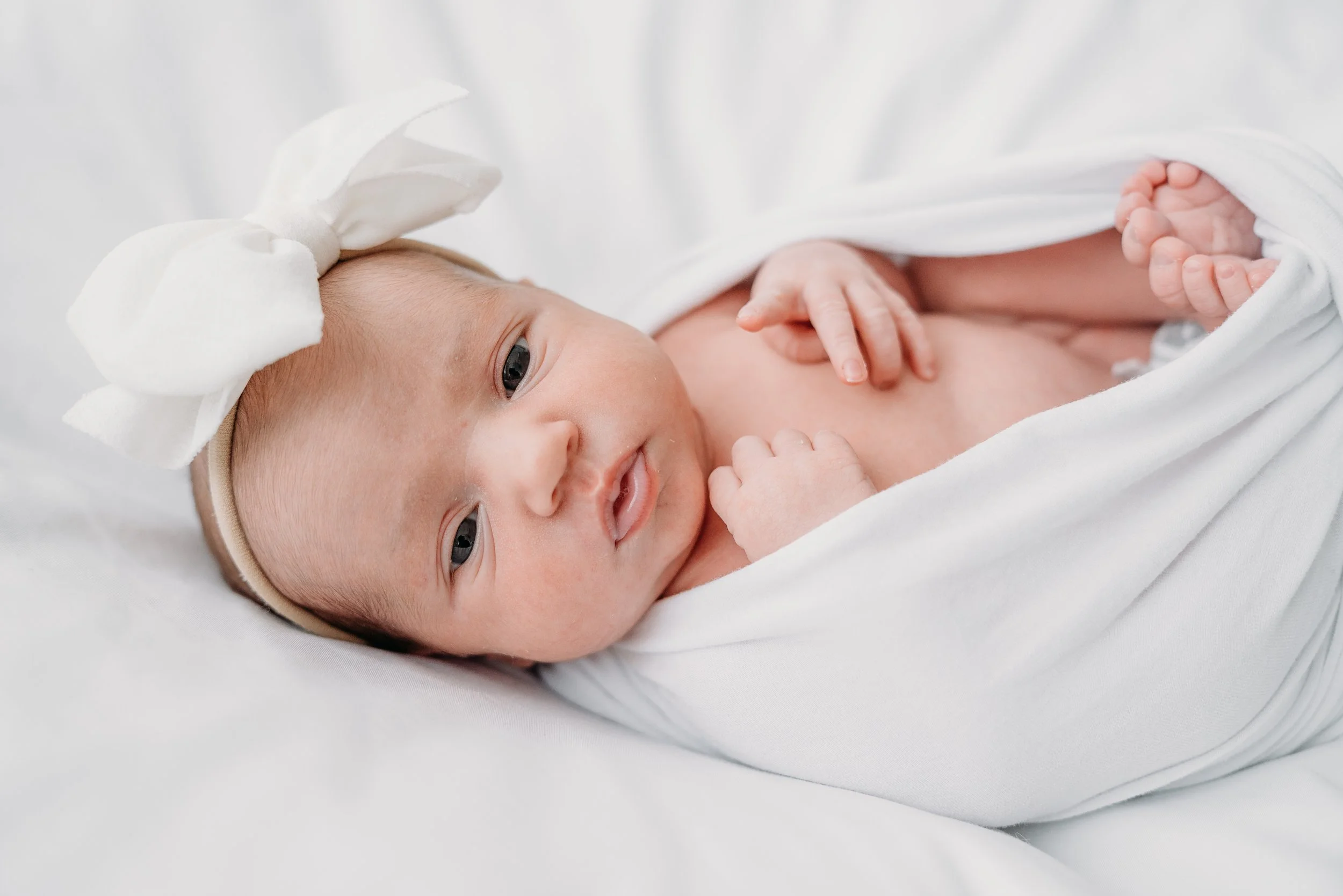 Cute newborn baby girl with a white bow headband swaddled in white blanket, lying on a white surface, looking at the camera.