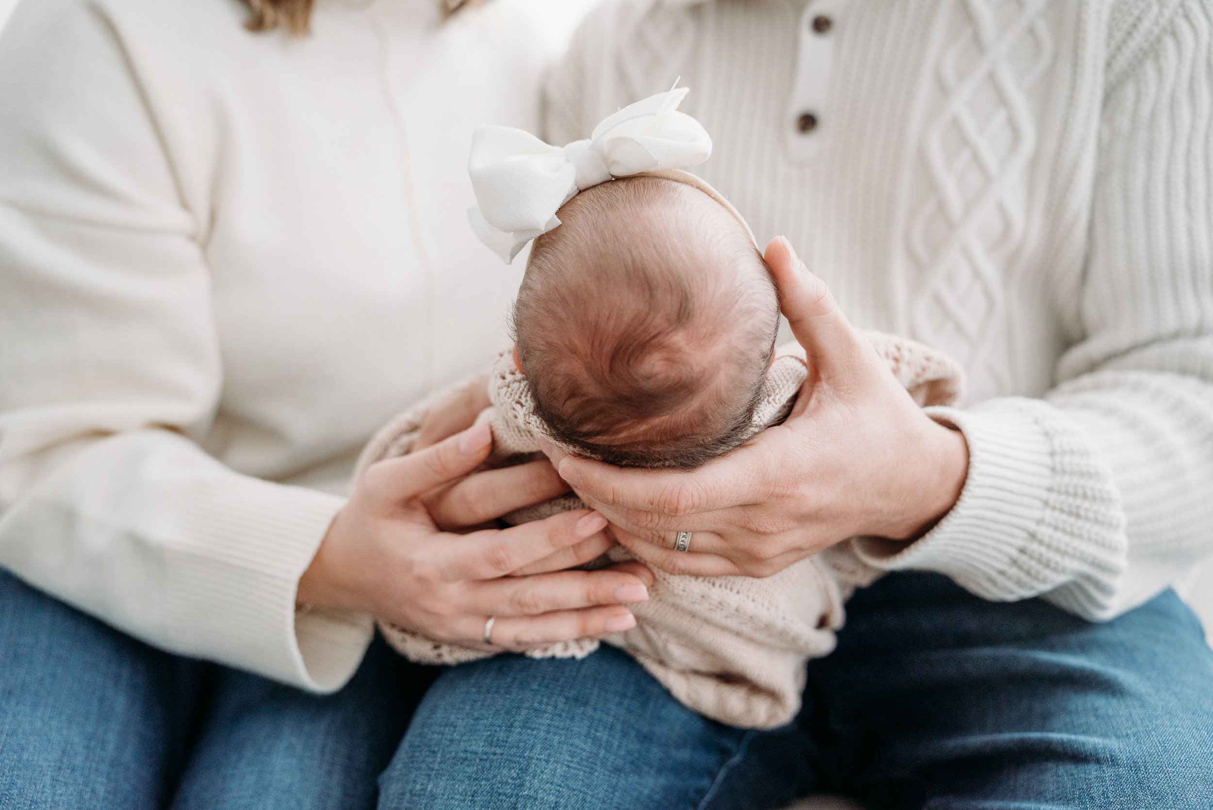 A person cradling a newborn baby on their lap, with the baby wearing a beige knitted outfit and a white bow headband, and the person wearing a cream-colored sweater.