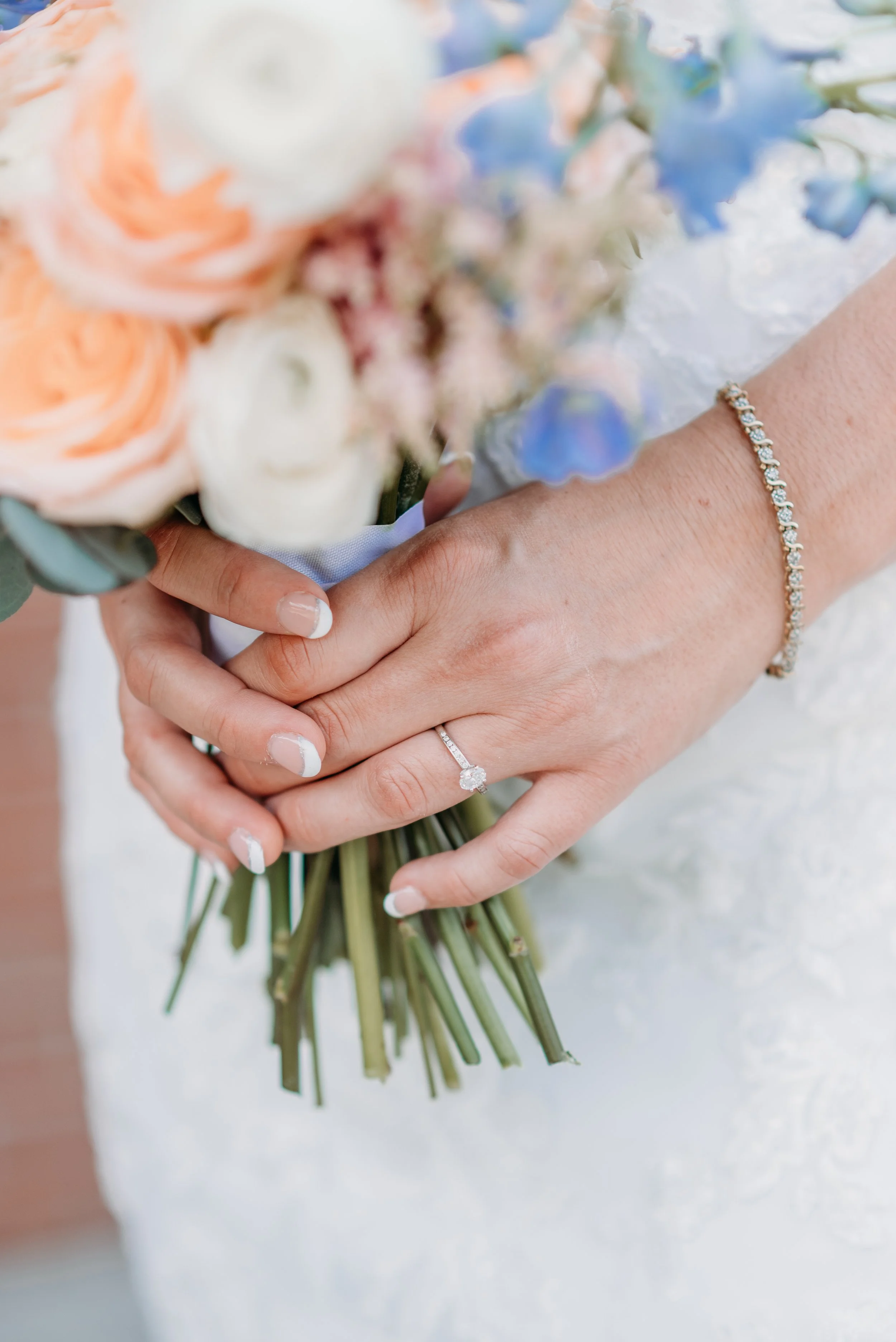 Close-up of a bride's hands holding a bouquet of flowers, showing an engagement ring, wedding band, and a bracelet.