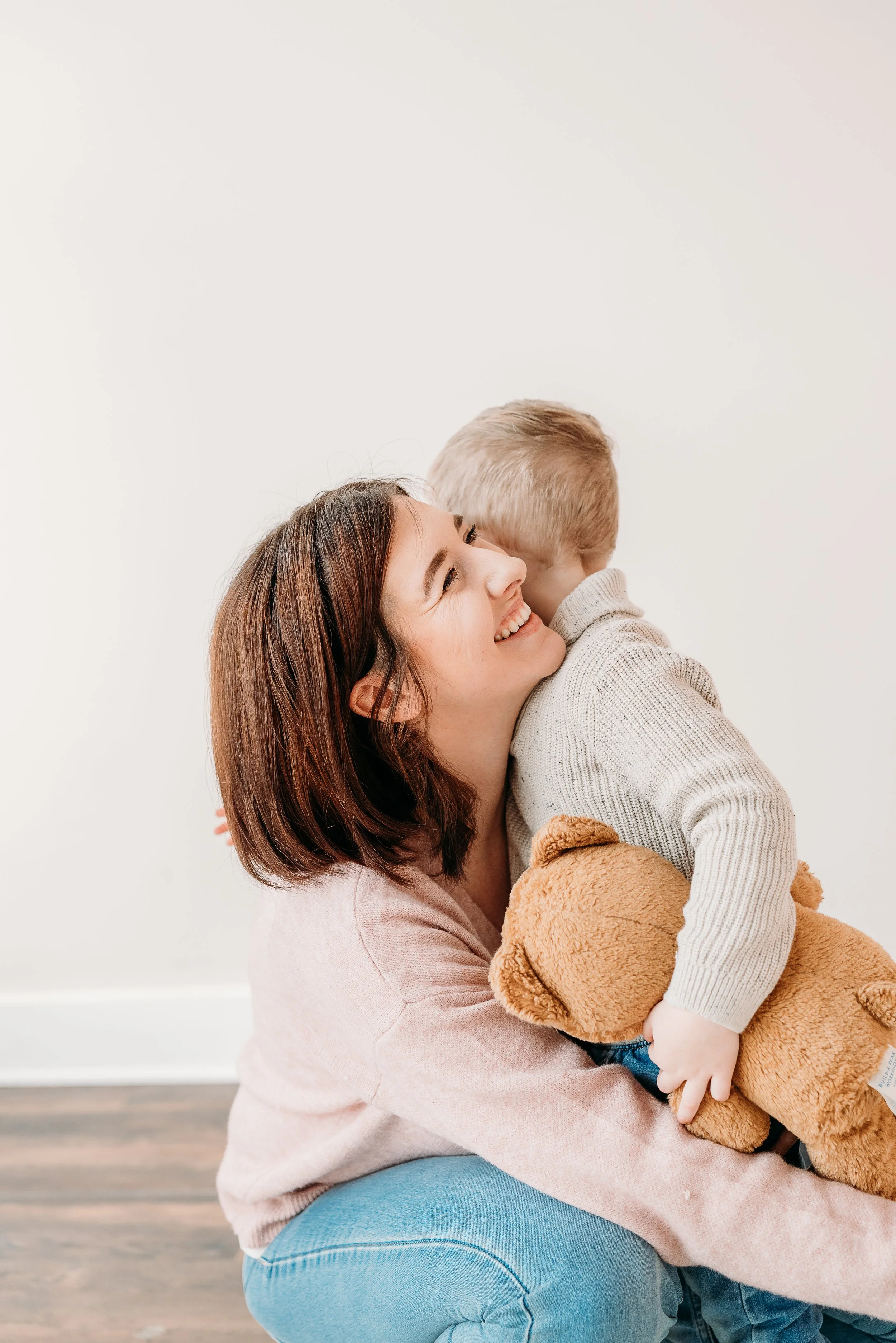 Woman hugging a young boy with a teddy bear, both smiling, against a plain white wall.