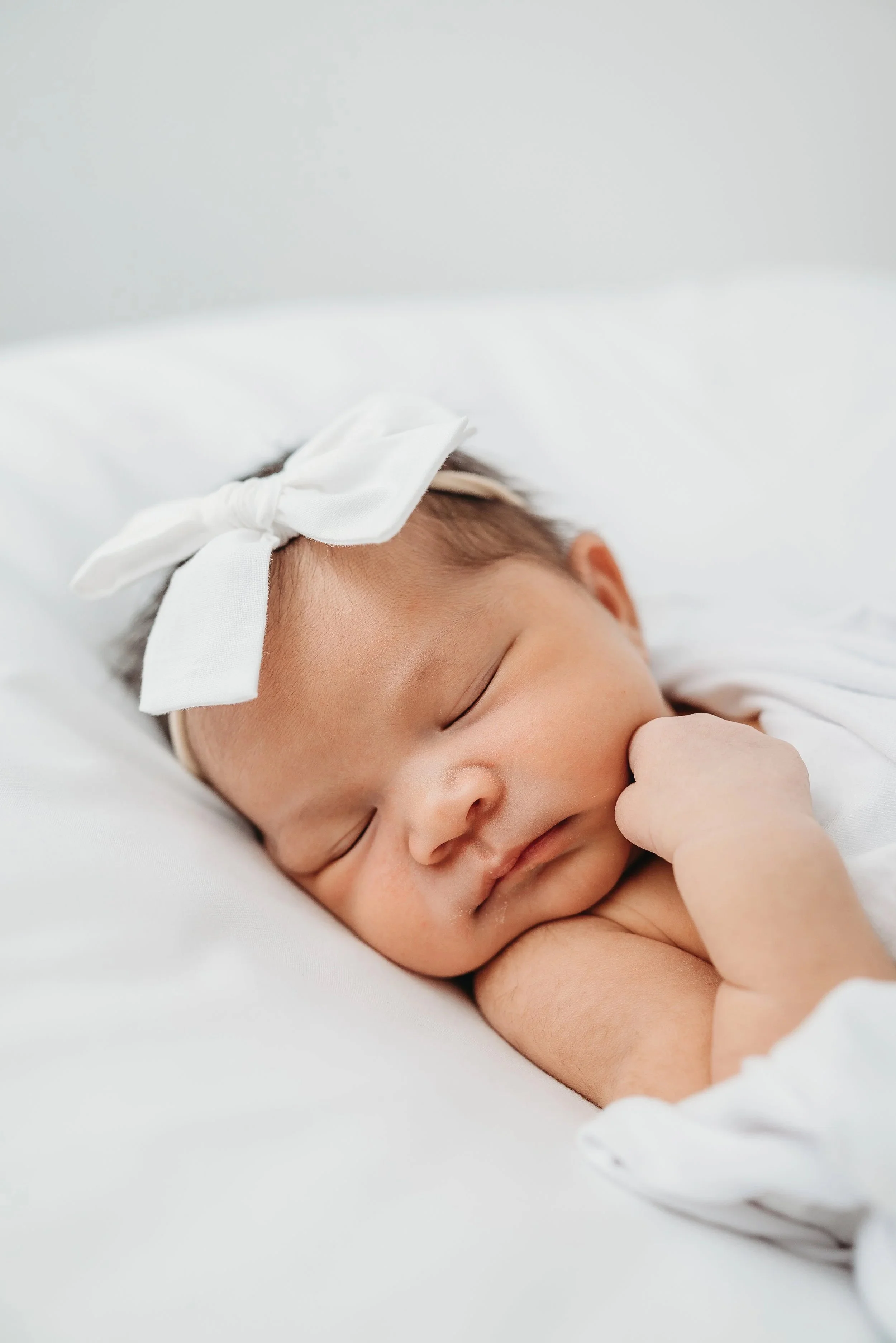 Close-up of a sleeping baby with a white bow headband, lying on a white bed.