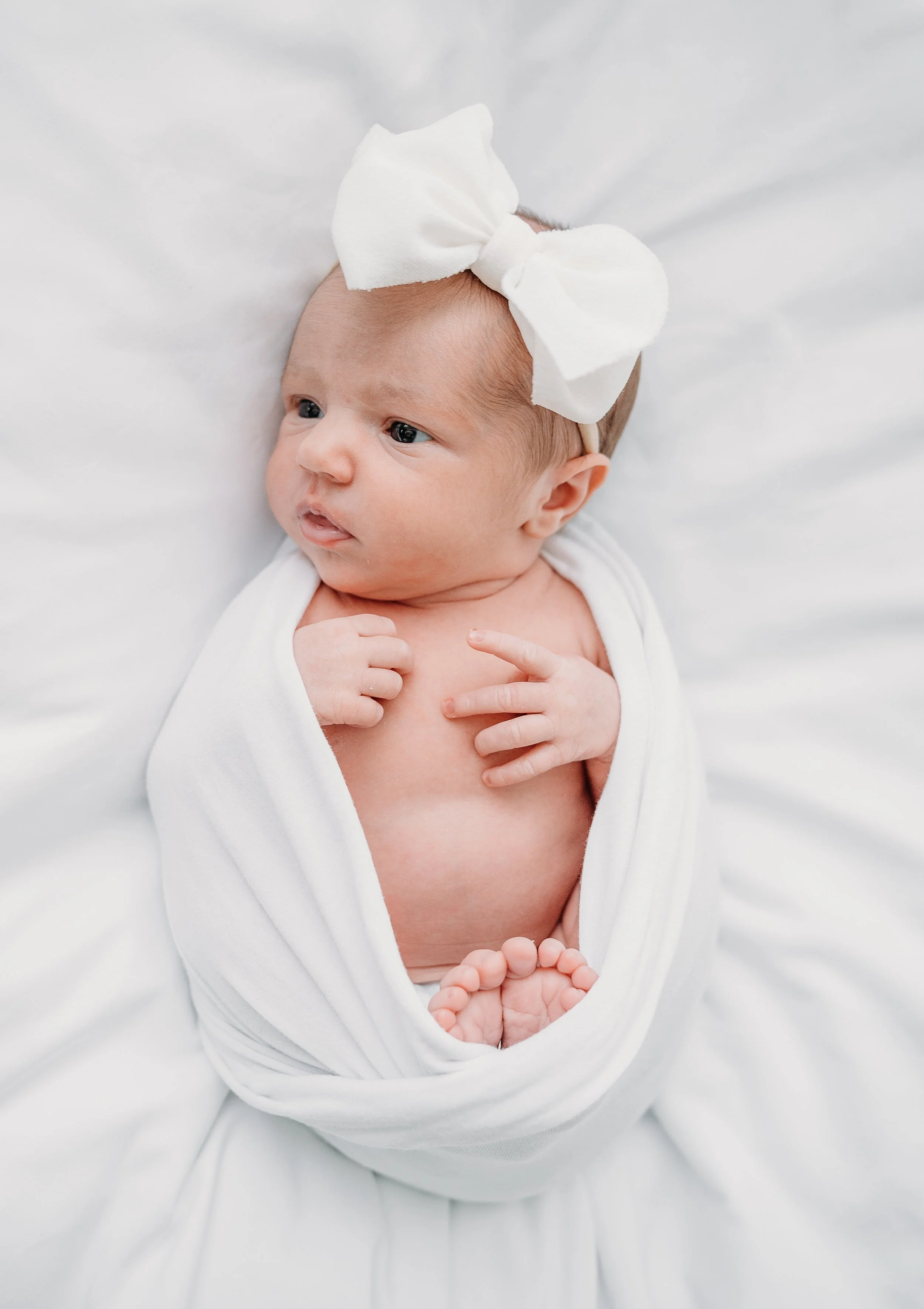 A newborn baby with a large white bow headband, wrapped in a white swaddle blanket, lying on a white surface.