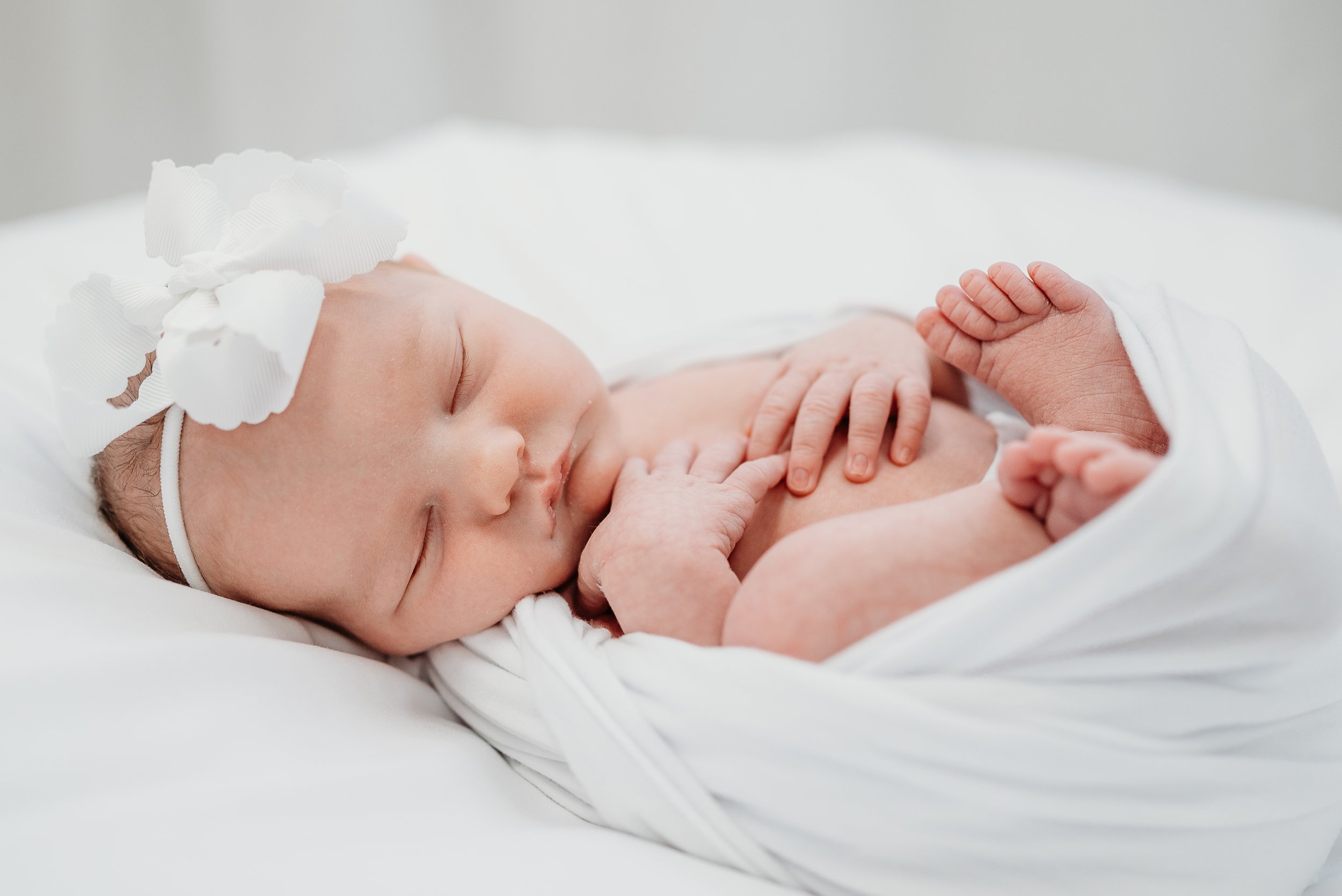 A sleeping newborn baby girl with a white bow on her head, lying on a white pillow and wrapped in a white blanket.