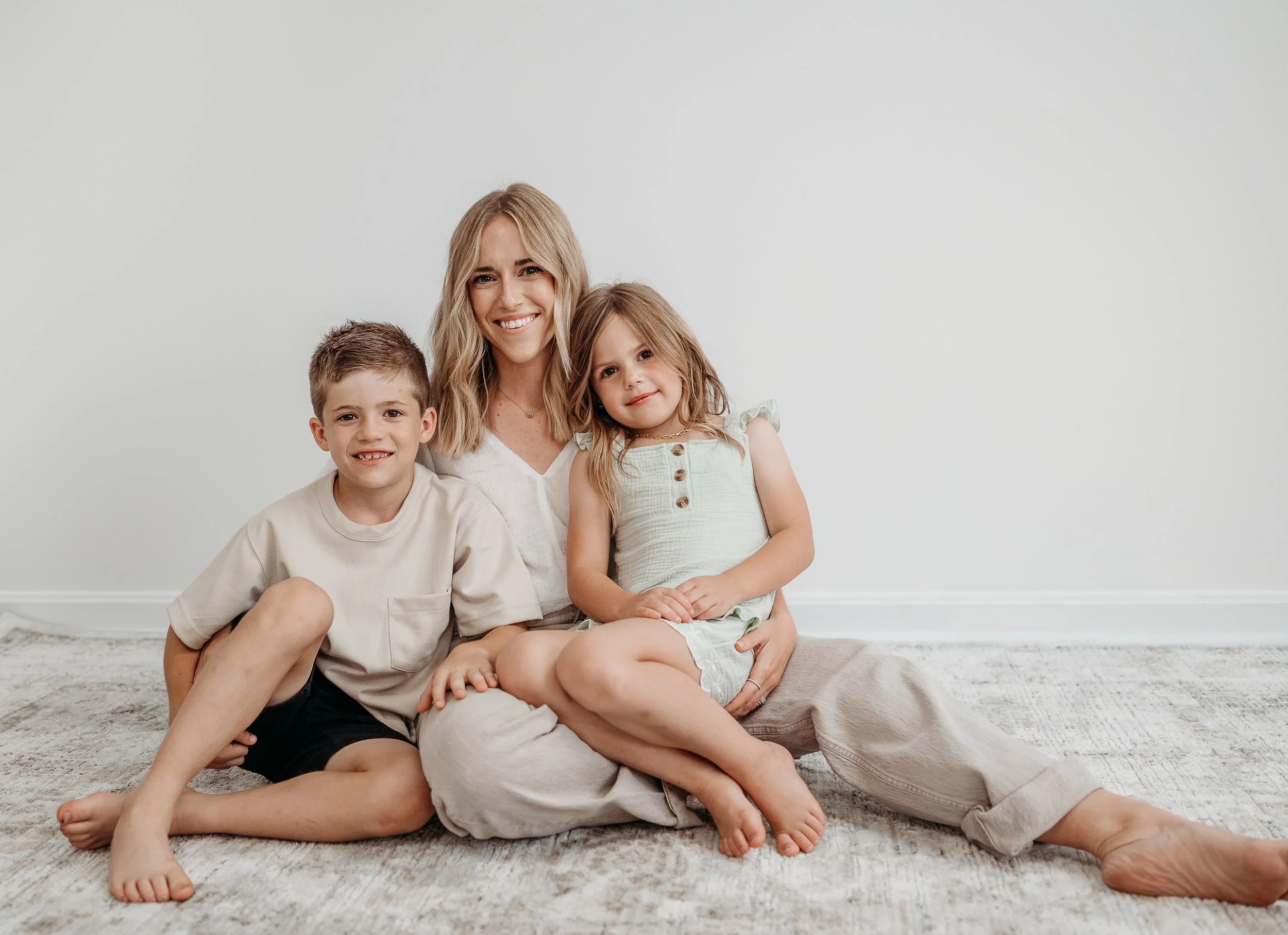 A woman sitting on the floor with a boy and a girl, all smiling and looking at the camera in a minimalistic room with white walls and a light-colored rug.