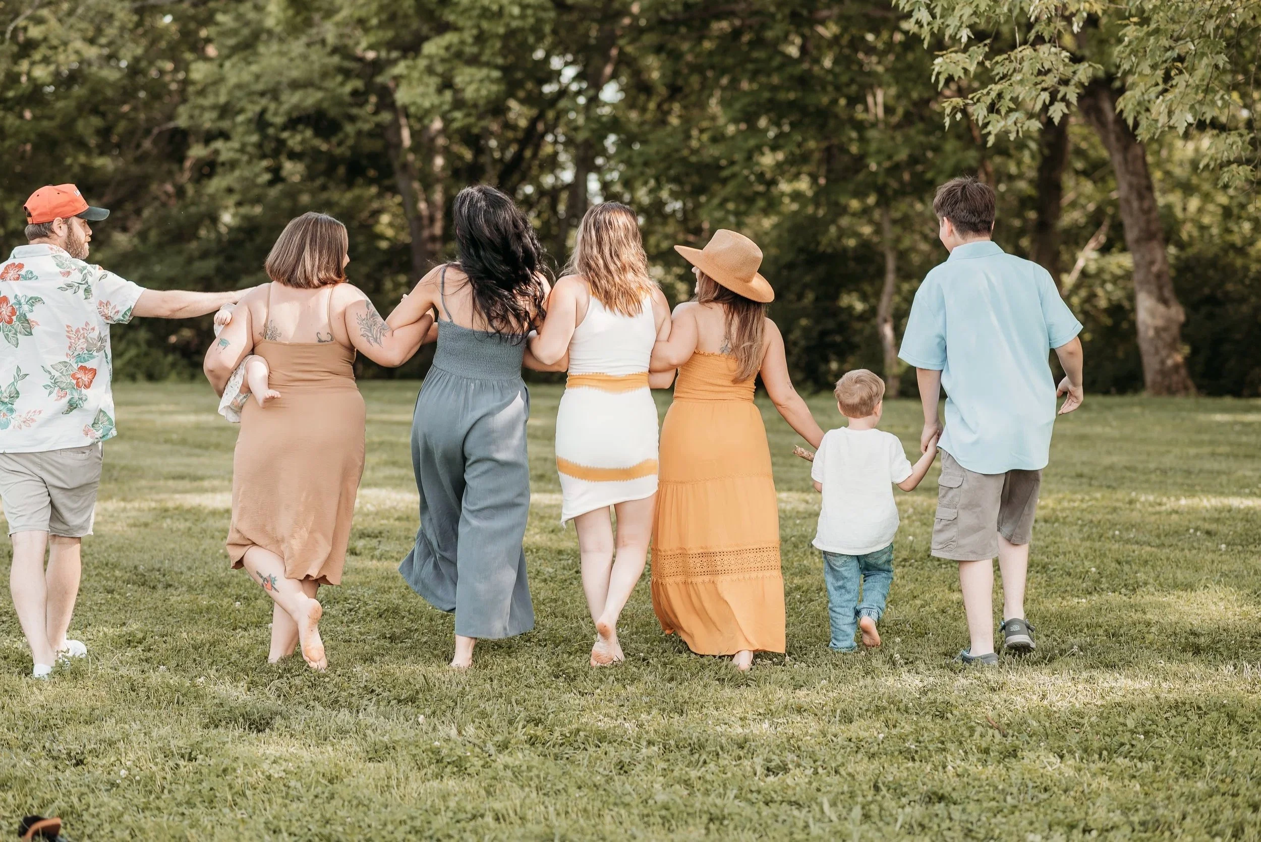 Group of people holding hands and walking in a park with green grass and trees in the background.