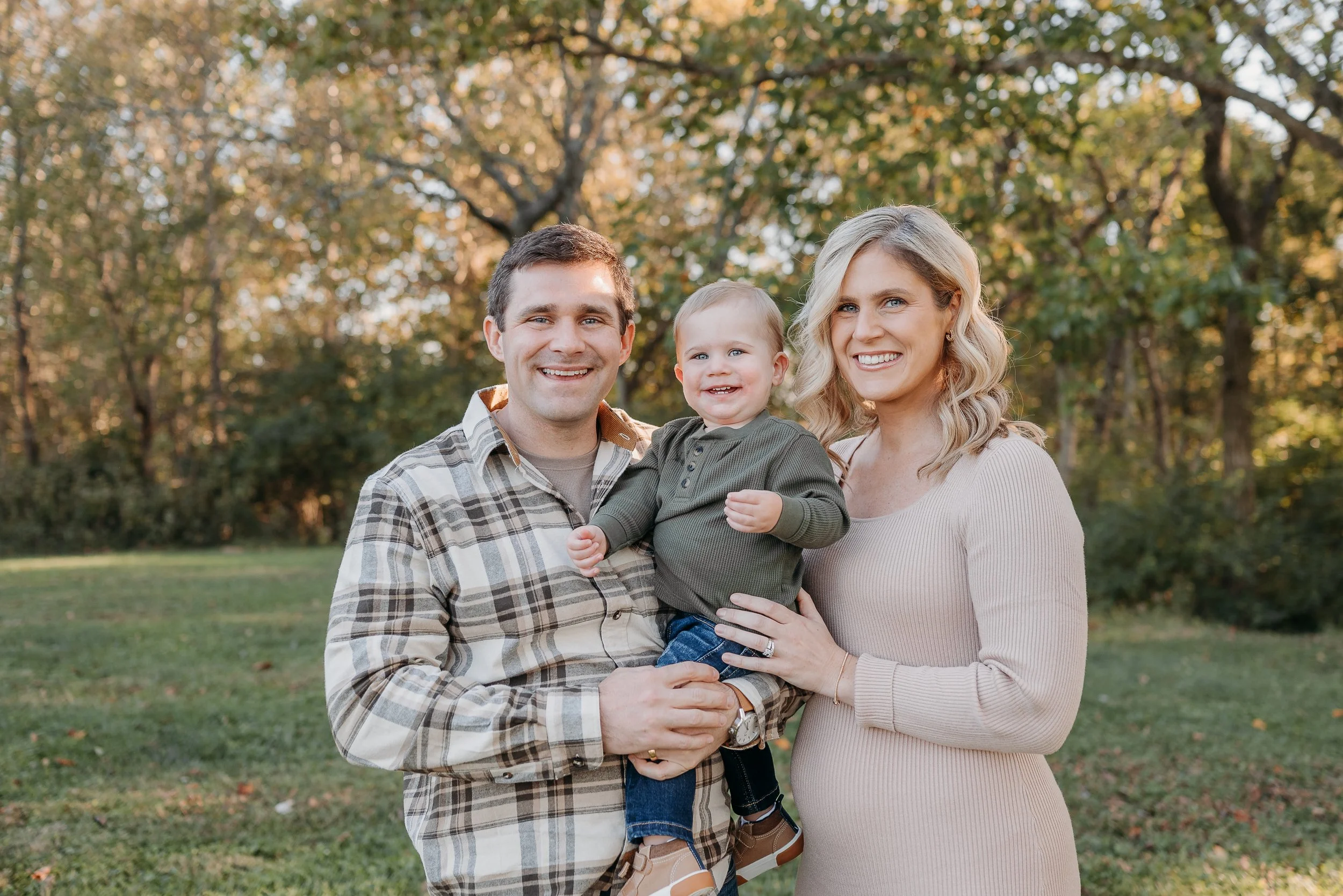 A happy family of three posing outdoors in a park during fall with trees in the background. The father is holding a smiling young boy, and the mother is standing next to them, all smiling.