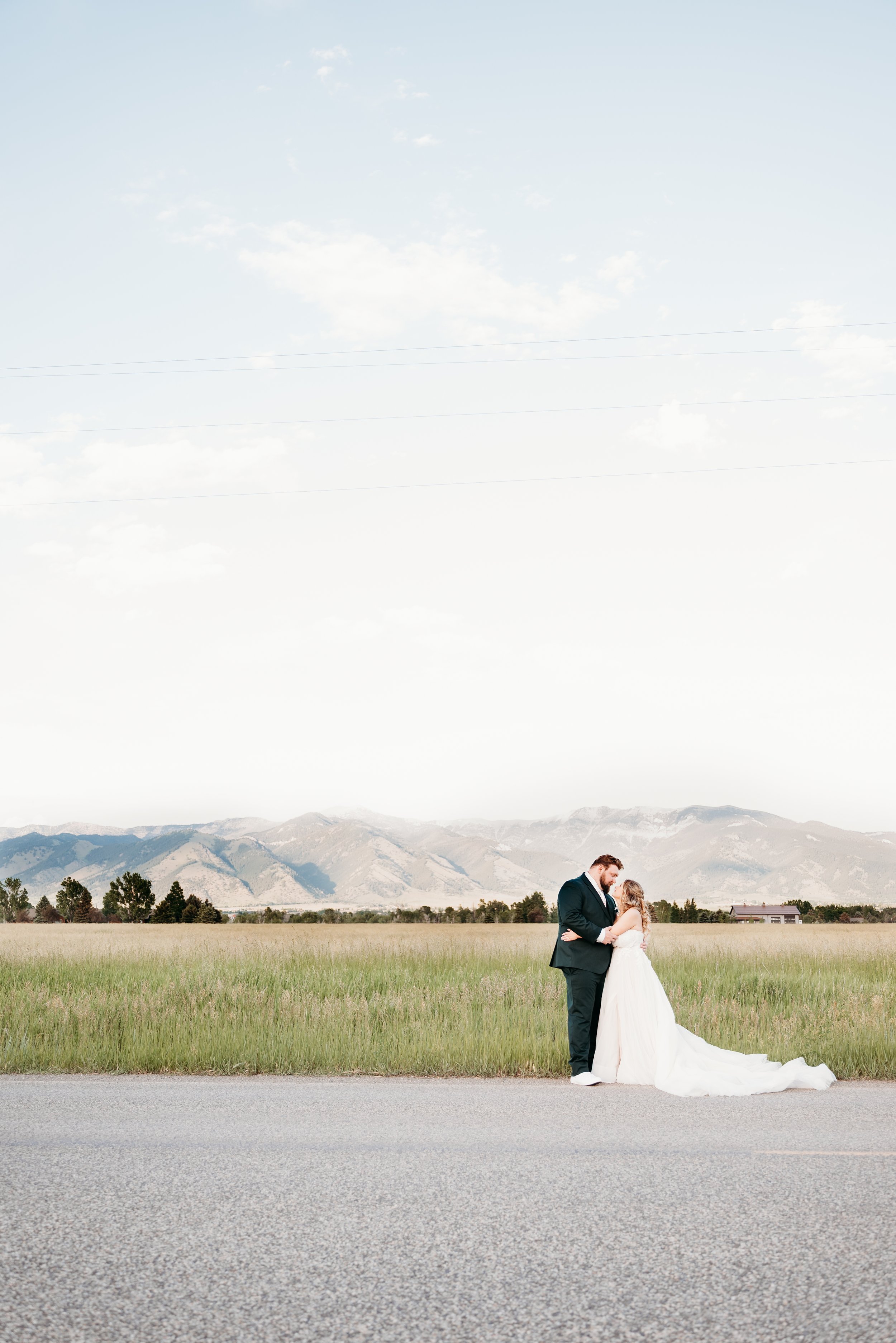 A bride and groom in wedding attire standing together on a roadside with mountains, trees, and a small building in the background.