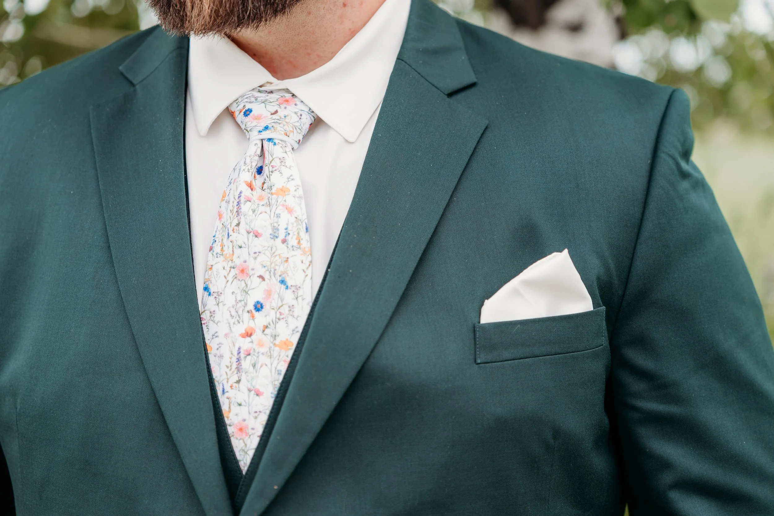 Close-up of a man wearing a dark green suit with a white shirt, a floral patterned tie, and a white pocket square, outdoors with blurred greenery in the background.