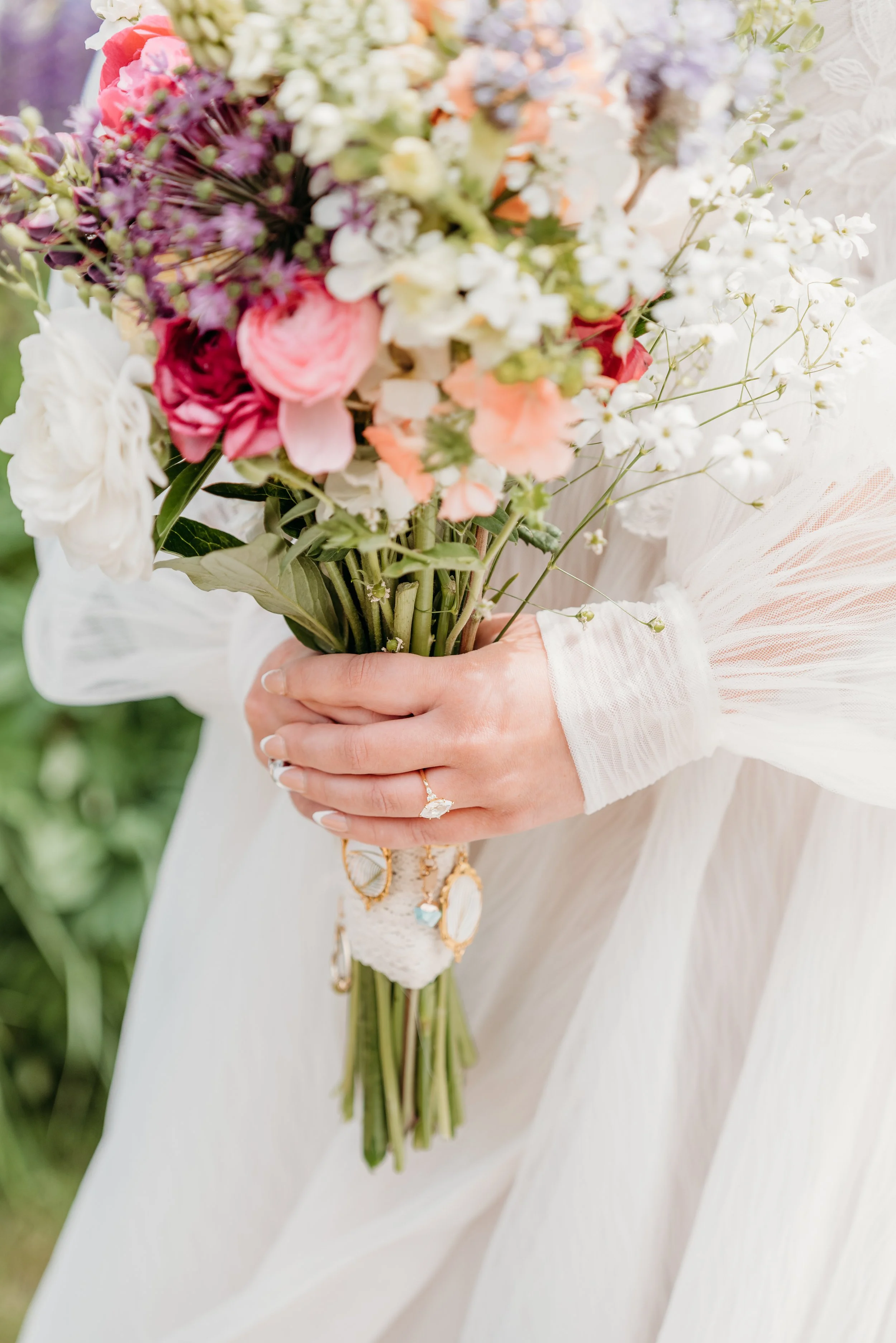 Close-up of a woman's hand holding a colorful bouquet of flowers, featuring white, pink, purple, and peach blossoms, with the woman wearing a white lace sleeve, wedding band, and bracelet, against a blurred green outdoor background.