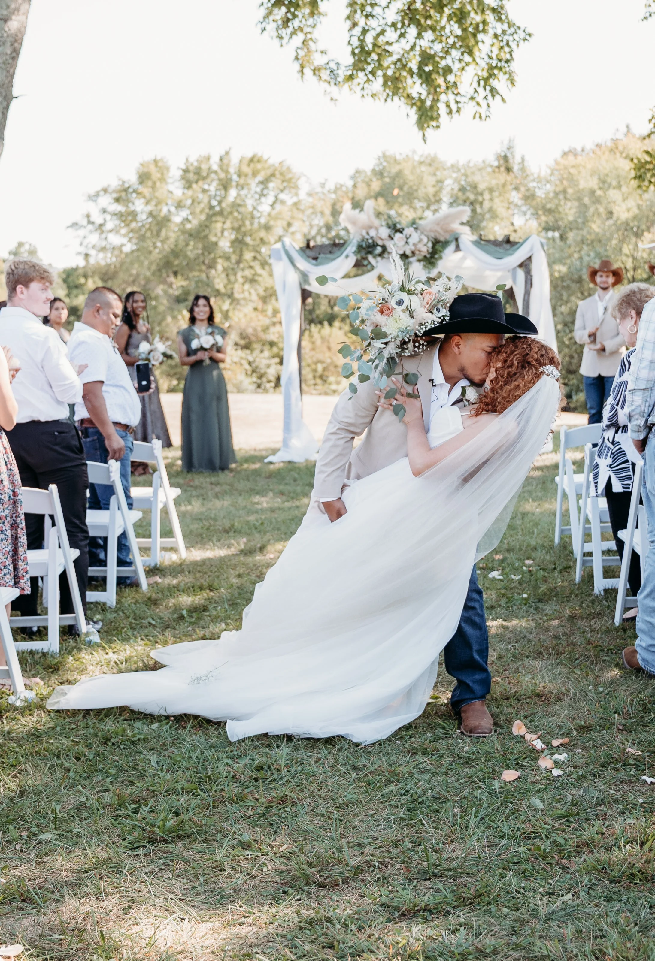 A bride and groom sharing a kiss during an outdoor wedding ceremony, with guests seated and standing around them, and a decorated arch in the background.